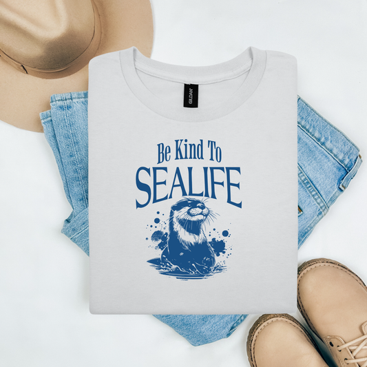 Gildan 5000 "Be Kind To Sealife" t shirt with a otter splashing in the water in a one color graphic. Picture on a flay lay mockup with pair of blue jeans, brown shoes and a hat accessory.