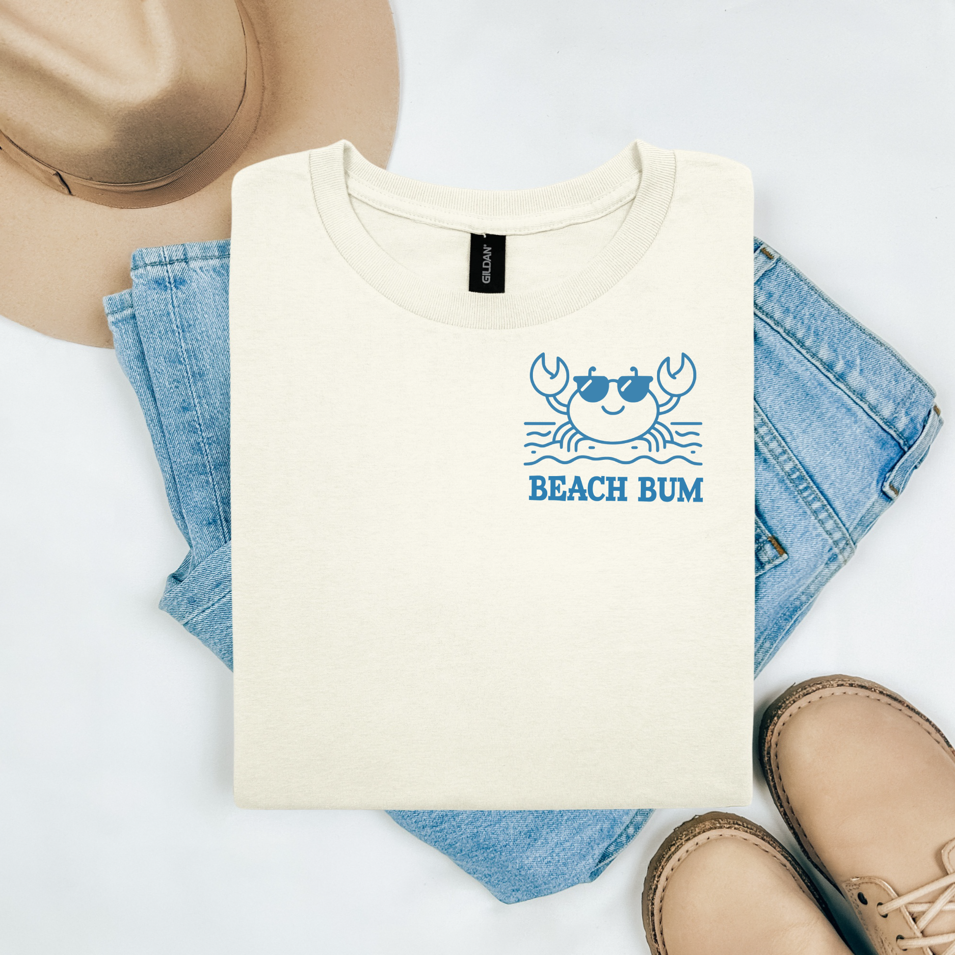 Gildan 5000 "Beach Bum" t shirt with a crab on the beach graphics in one color. Picture on a flay lay mockup with pair of blue jeans, brown shoes and a hat accessory.