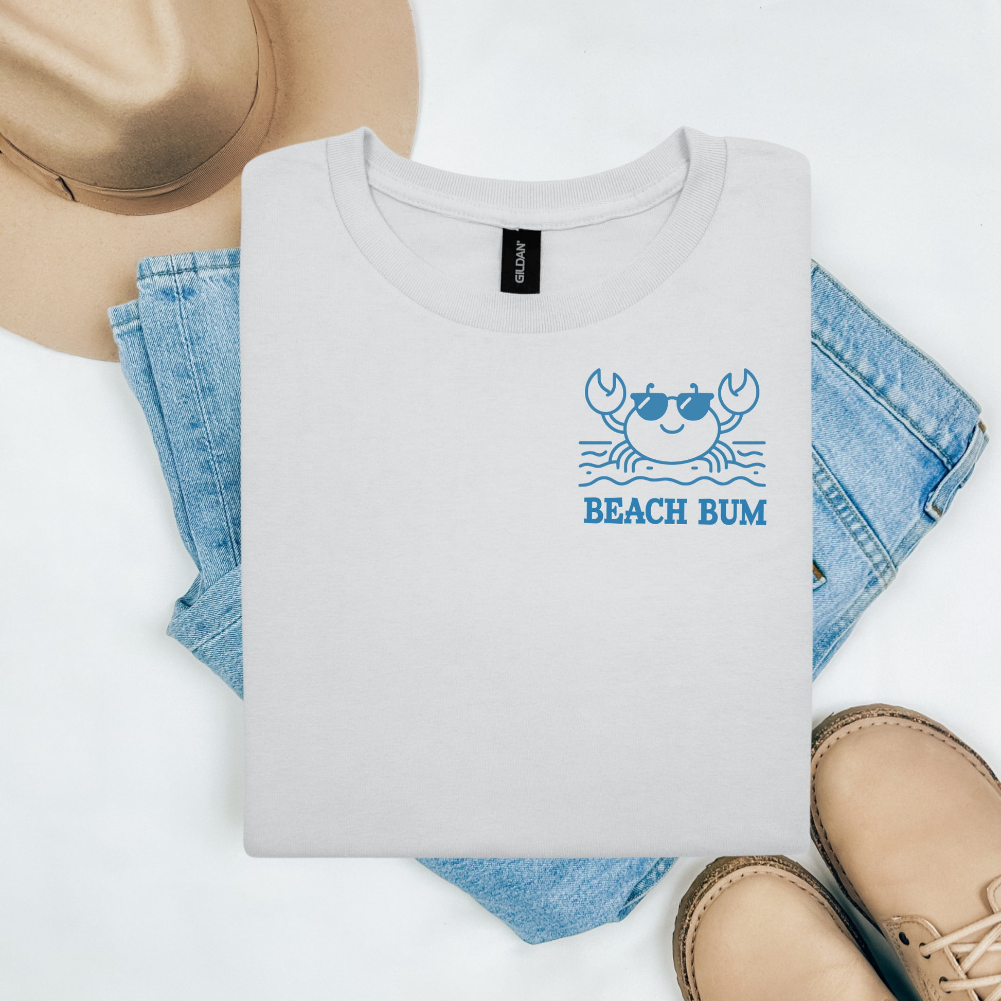 Gildan 5000 "Beach Bum" t shirt with a crab on the beach graphics in one color. Picture on a flay lay mockup with pair of blue jeans, brown shoes and a hat accessory.