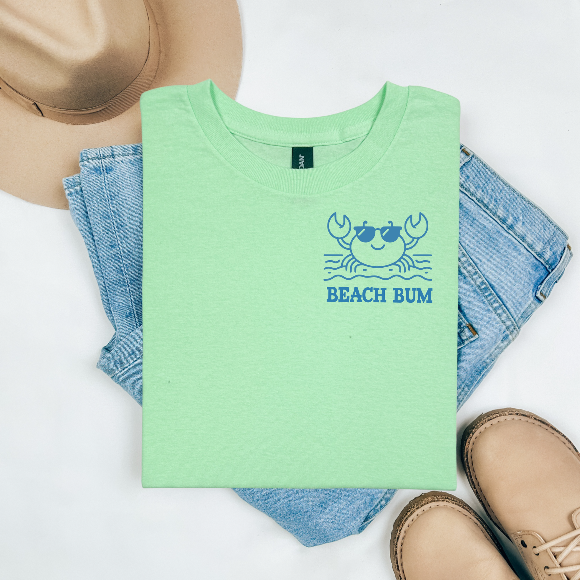 Gildan 5000 "Beach Bum" t shirt with a crab on the beach graphics in one color. Picture on a flay lay mockup with pair of blue jeans, brown shoes and a hat accessory.