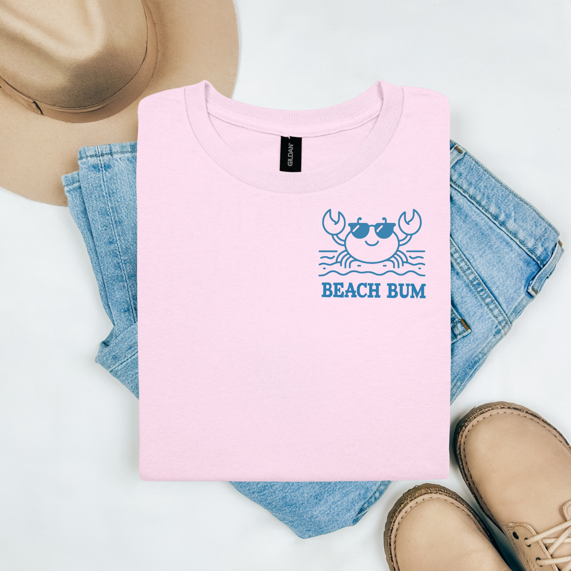 Gildan 5000 "Beach Bum" t shirt with a crab on the beach graphics in one color. Picture on a flay lay mockup with pair of blue jeans, brown shoes and a hat accessory.
