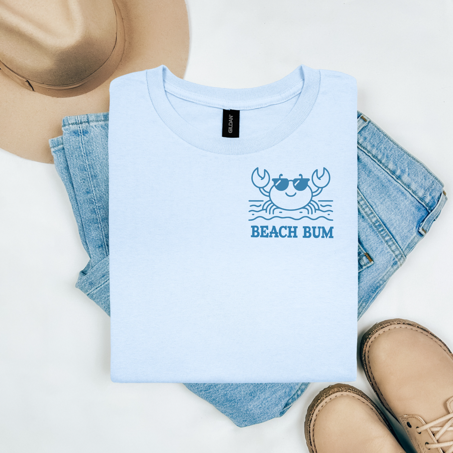 Gildan 5000 "Beach Bum" t shirt with a crab on the beach graphics in one color. Picture on a flay lay mockup with pair of blue jeans, brown shoes and a hat accessory.