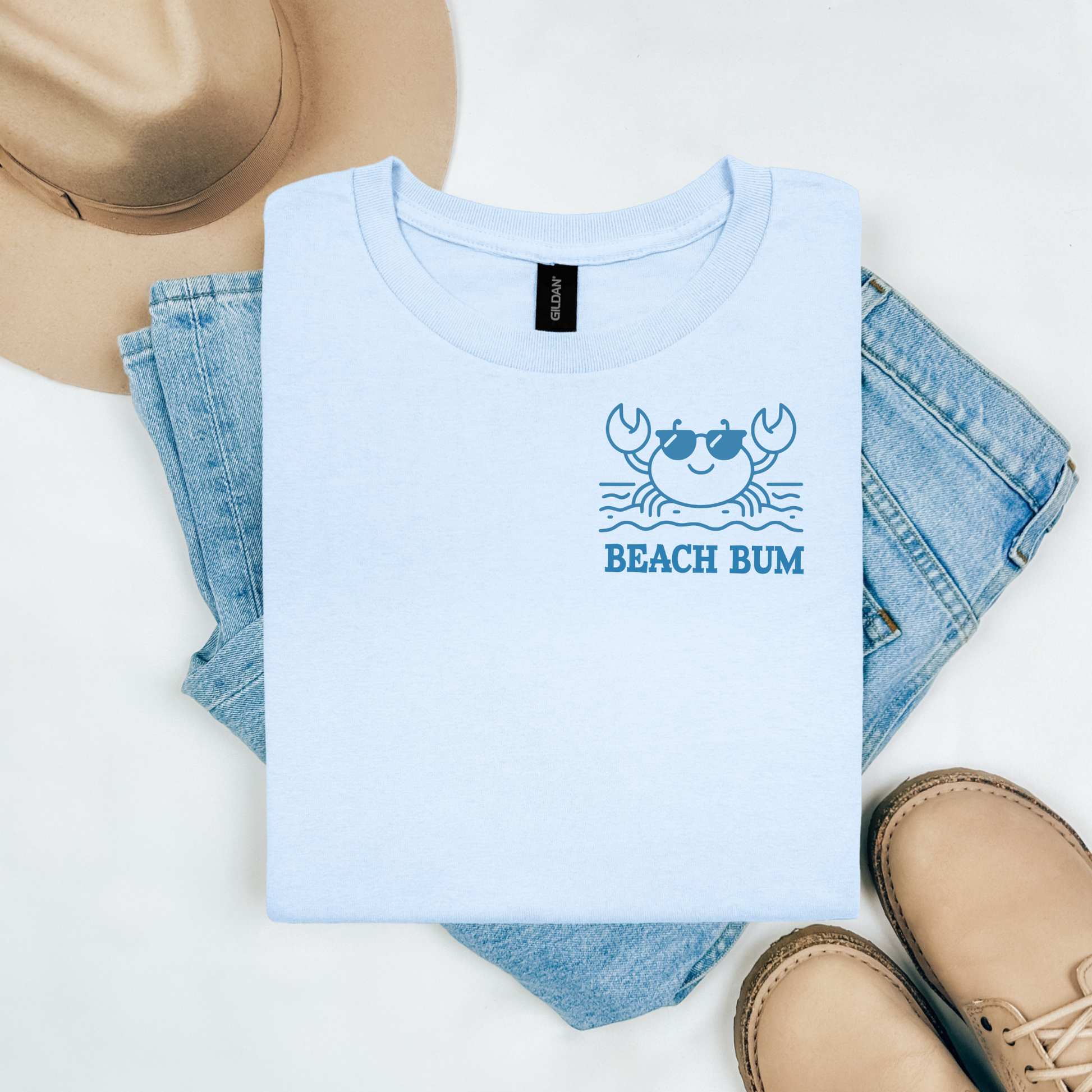 Gildan 5000 "Beach Bum" t shirt with a crab on the beach graphics in one color. Picture on a flay lay mockup with pair of blue jeans, brown shoes and a hat accessory.