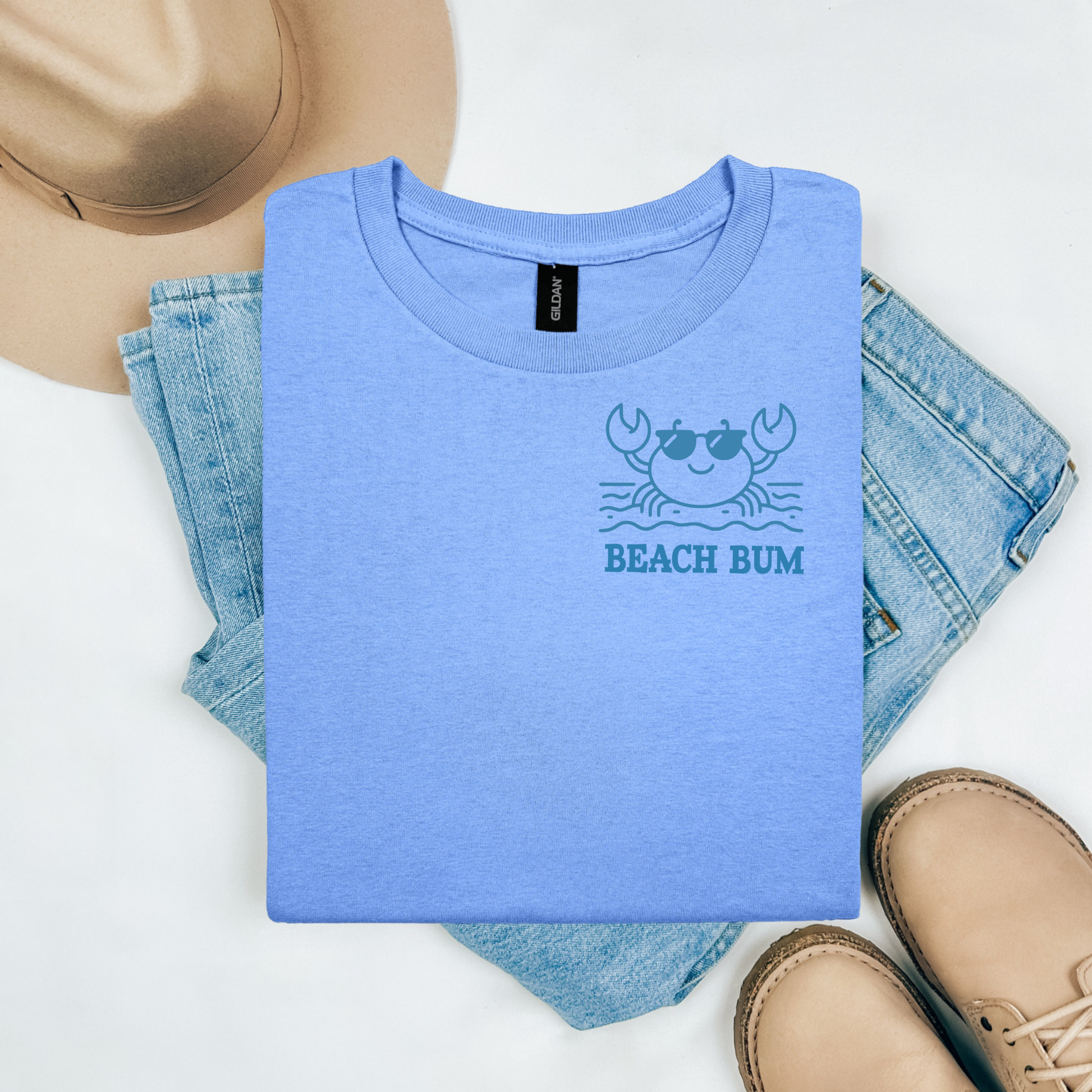 Gildan 5000 "Beach Bum" t shirt with a crab on the beach graphics in one color. Picture on a flay lay mockup with pair of blue jeans, brown shoes and a hat accessory.