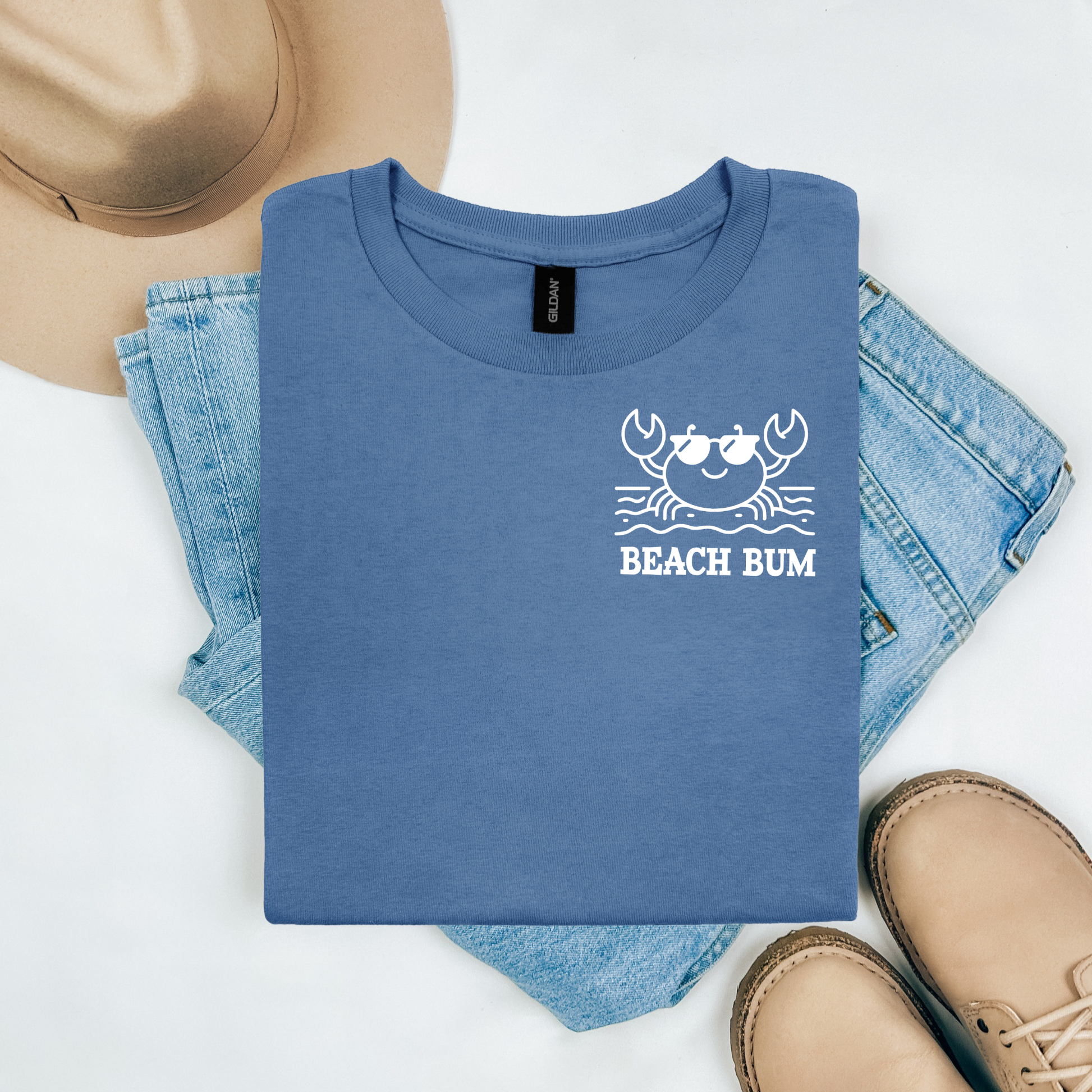 Gildan 5000 "Beach Bum" t shirt with a crab on the beach graphics in one color. Picture on a flay lay mockup with pair of blue jeans, brown shoes and a hat accessory.