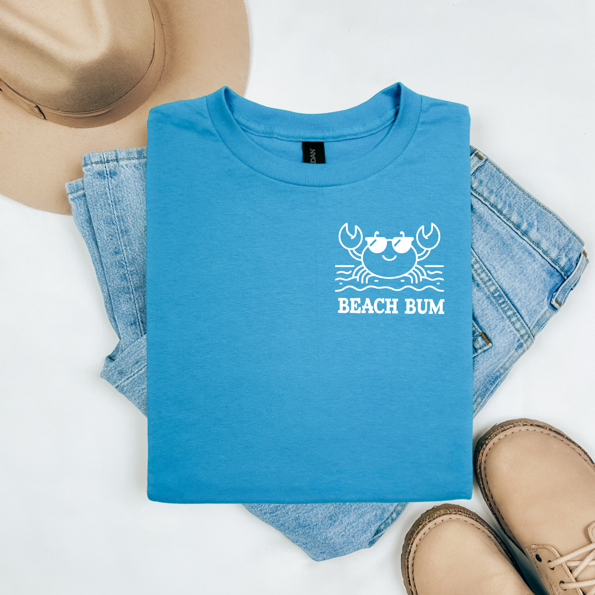 Gildan 5000 "Beach Bum" t shirt with a crab on the beach graphics in one color. Picture on a flay lay mockup with pair of blue jeans, brown shoes and a hat accessory.