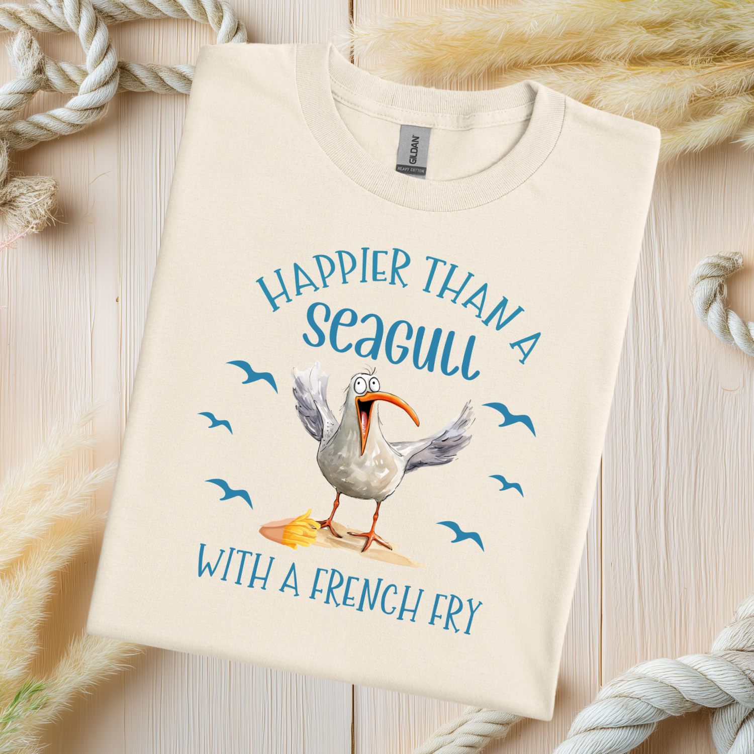 Beige t-shirt with a seagull graphic and text on a wooden surface with ropes.