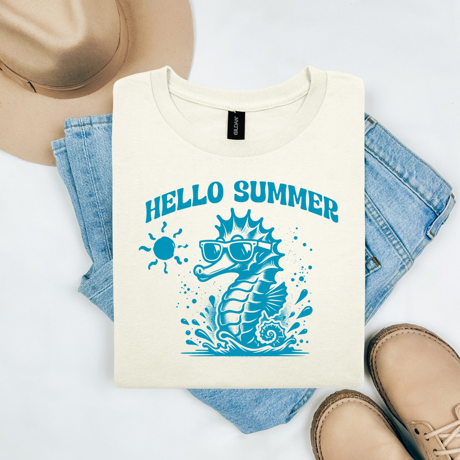 Gildan 5000 "Hello Summer" t shirt with a cute seahorse with sunglasses in one color graphics. Picture on a flay lay mockup with pair of blue jeans, brown shoes and a hat accessory.