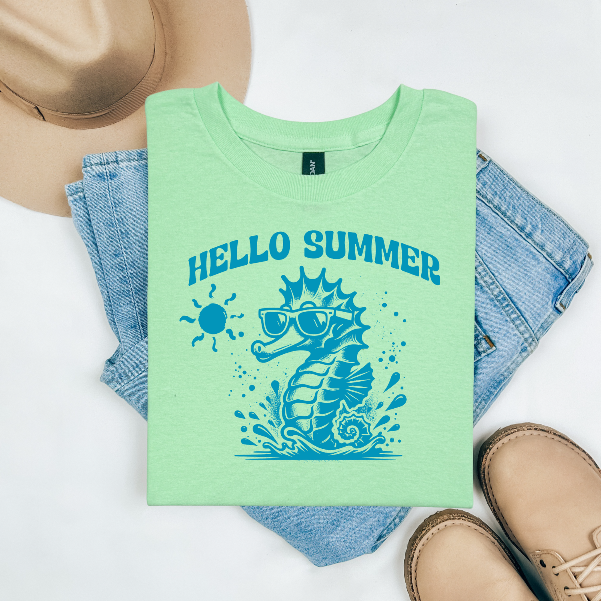 Gildan 5000 "Hello Summer" t shirt with a cute seahorse with sunglasses in one color graphics. Picture on a flay lay mockup with pair of blue jeans, brown shoes and a hat accessory.