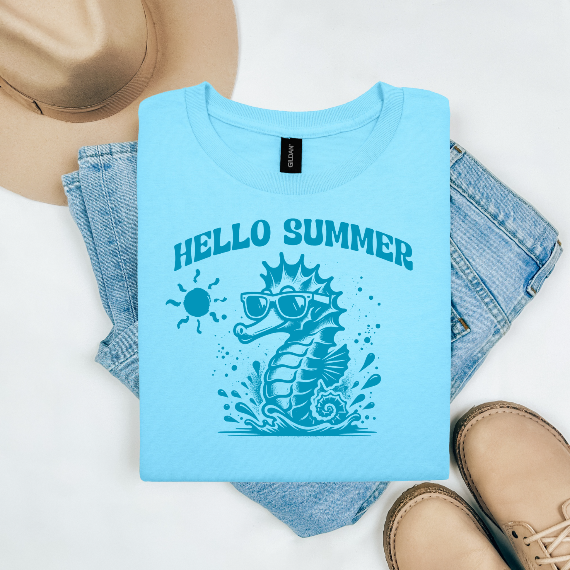 Gildan 5000 "Hello Summer" t shirt with a cute seahorse with sunglasses in one color graphics. Picture on a flay lay mockup with pair of blue jeans, brown shoes and a hat accessory.