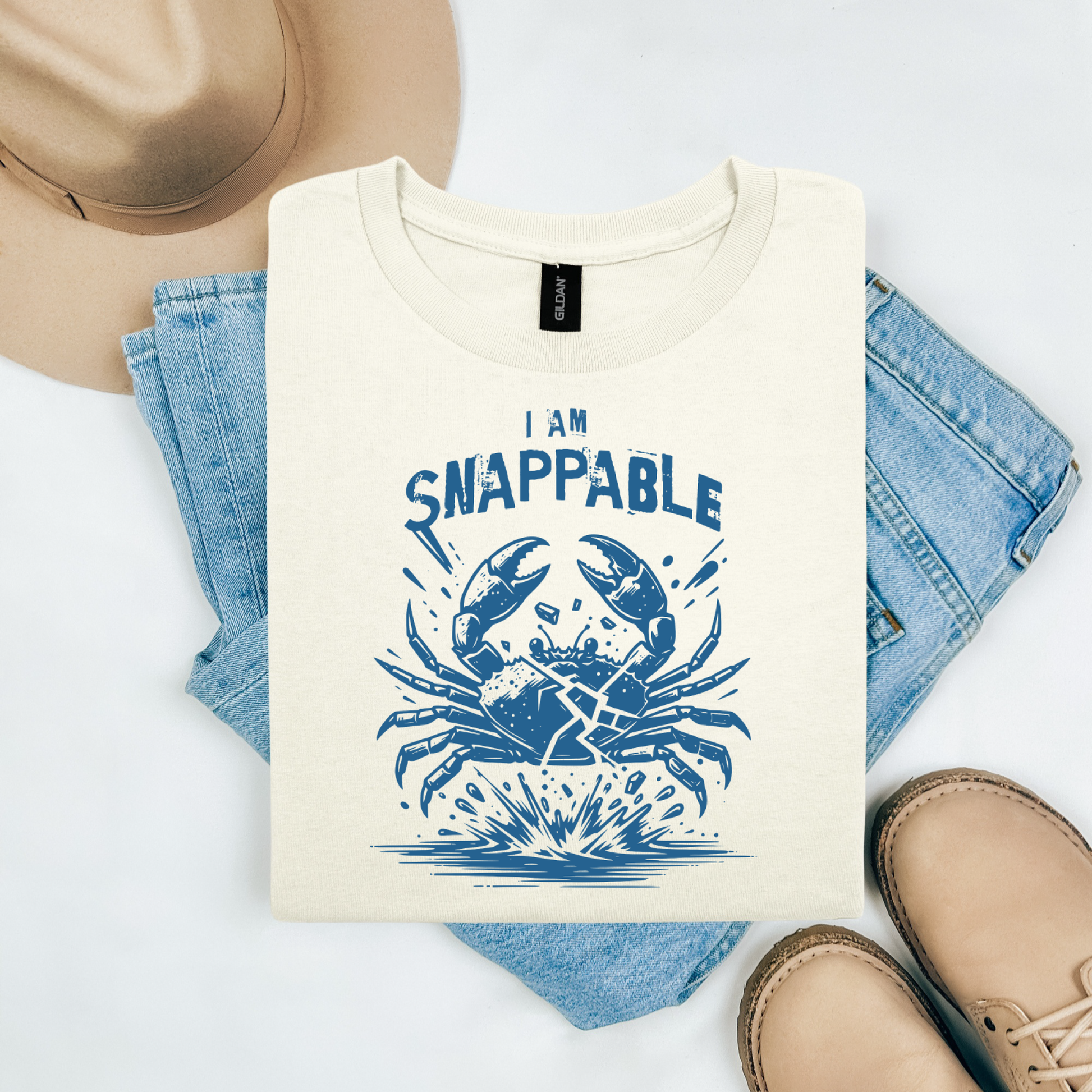 Gildan 5000 "I Am Snappable" t shirt with a crab cracked in half in a one color graphic. Picture on a flay lay mockup with pair of blue jeans, brown shoes and a hat accessory.