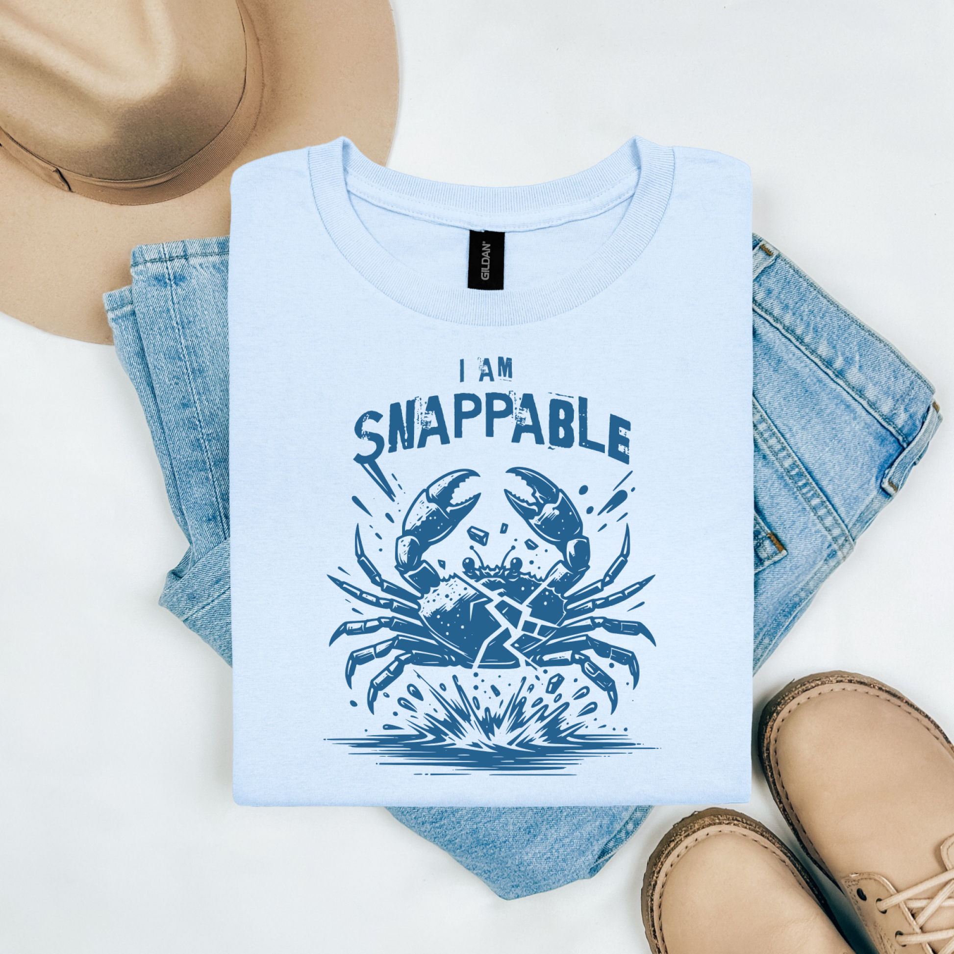 Gildan 5000 "I Am Snappable" t shirt with a crab cracked in half in a one color graphic. Picture on a flay lay mockup with pair of blue jeans, brown shoes and a hat accessory.