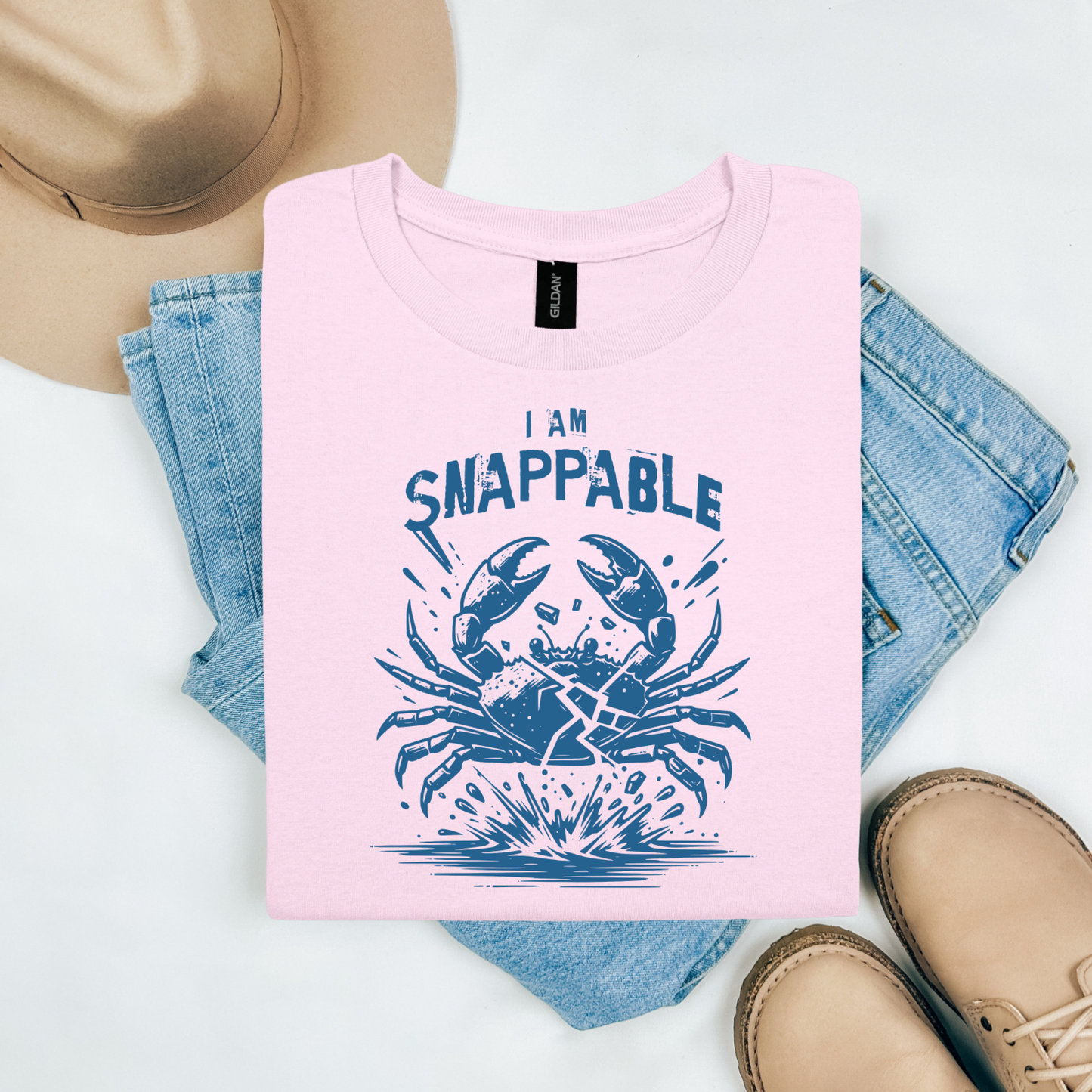 Gildan 5000 "I Am Snappable" t shirt with a crab cracked in half in a one color graphic. Picture on a flay lay mockup with pair of blue jeans, brown shoes and a hat accessory.