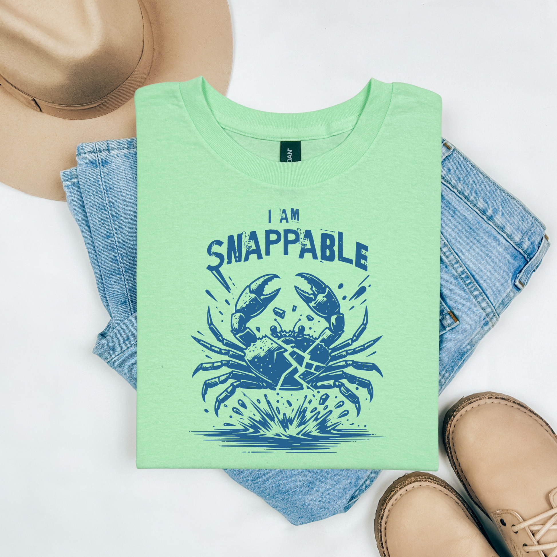 Gildan 5000 "I Am Snappable" t shirt with a crab cracked in half in a one color graphic. Picture on a flay lay mockup with pair of blue jeans, brown shoes and a hat accessory.
