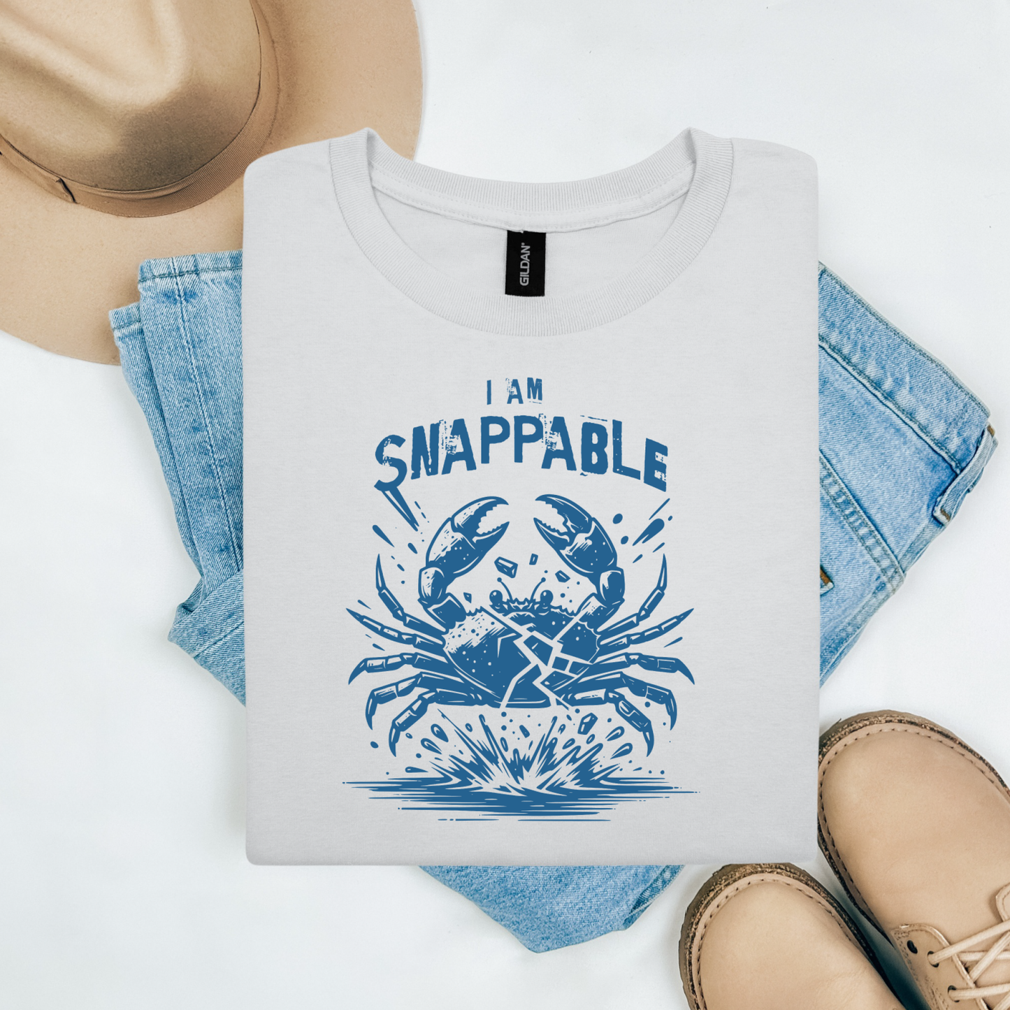Gildan 5000 "I Am Snappable" t shirt with a crab cracked in half in a one color graphic. Picture on a flay lay mockup with pair of blue jeans, brown shoes and a hat accessory.