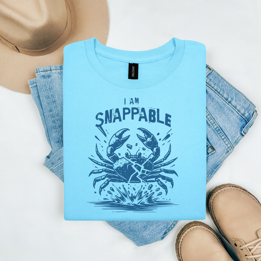 Gildan 5000 "I Am Snappable" t shirt with a crab cracked in half in a one color graphic. Picture on a flay lay mockup with pair of blue jeans, brown shoes and a hat accessory.