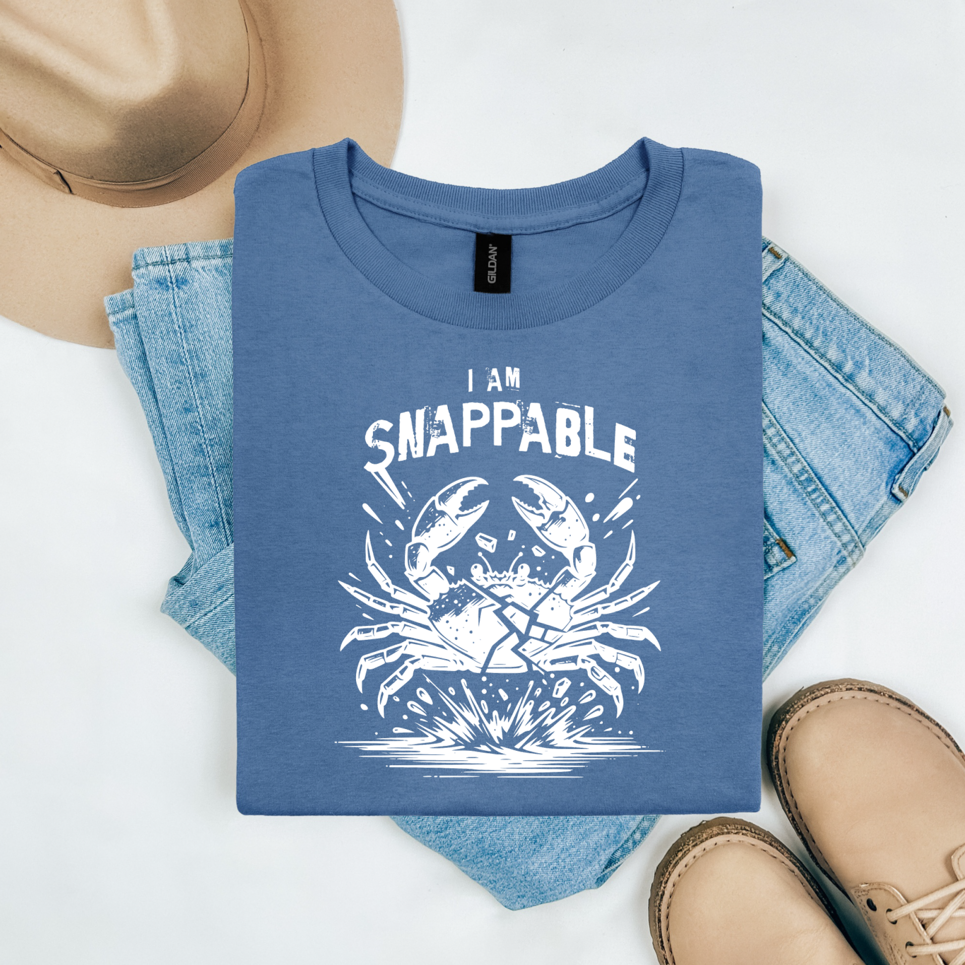 Gildan 5000 "I Am Snappable" t shirt with a crab cracked in half in a one color graphic. Picture on a flay lay mockup with pair of blue jeans, brown shoes and a hat accessory.