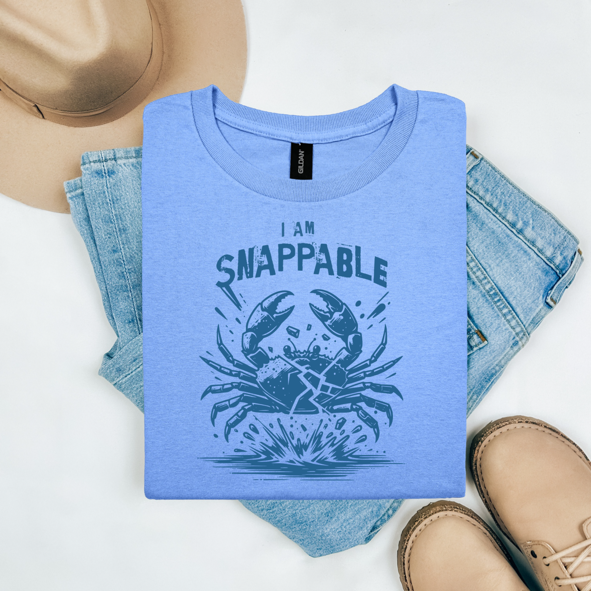 Gildan 5000 "I Am Snappable" t shirt with a crab cracked in half in a one color graphic. Picture on a flay lay mockup with pair of blue jeans, brown shoes and a hat accessory.