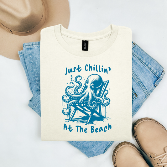 Gildan 5000 "Just Chillin' At The Beach" t shirt with an octopus setting in a beach chair in one color graphics. Picture on a flay lay mockup with pair of blue jeans, brown shoes and a hat accessory.