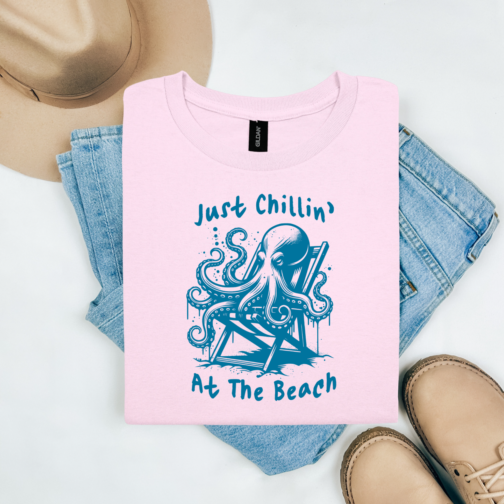 Gildan 5000 "Just Chillin' At The Beach" t shirt with an octopus setting in a beach chair in one color graphics. Picture on a flay lay mockup with pair of blue jeans, brown shoes and a hat accessory.