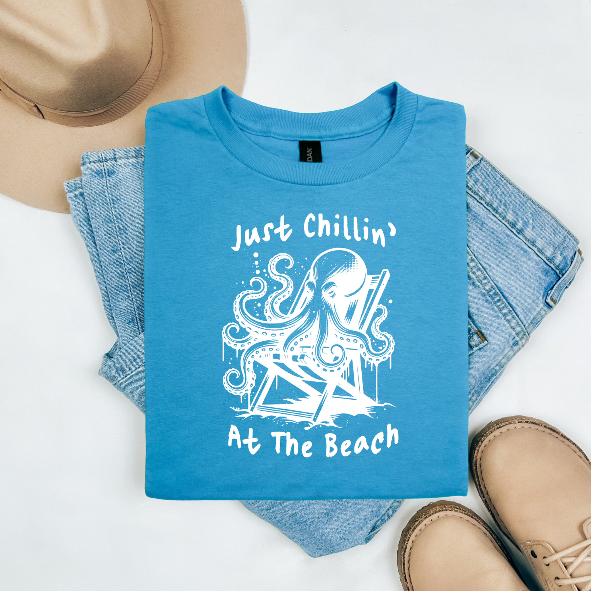 Gildan 5000 "Just Chillin' At The Beach" t shirt with an octopus setting in a beach chair in one color graphics. Picture on a flay lay mockup with pair of blue jeans, brown shoes and a hat accessory.