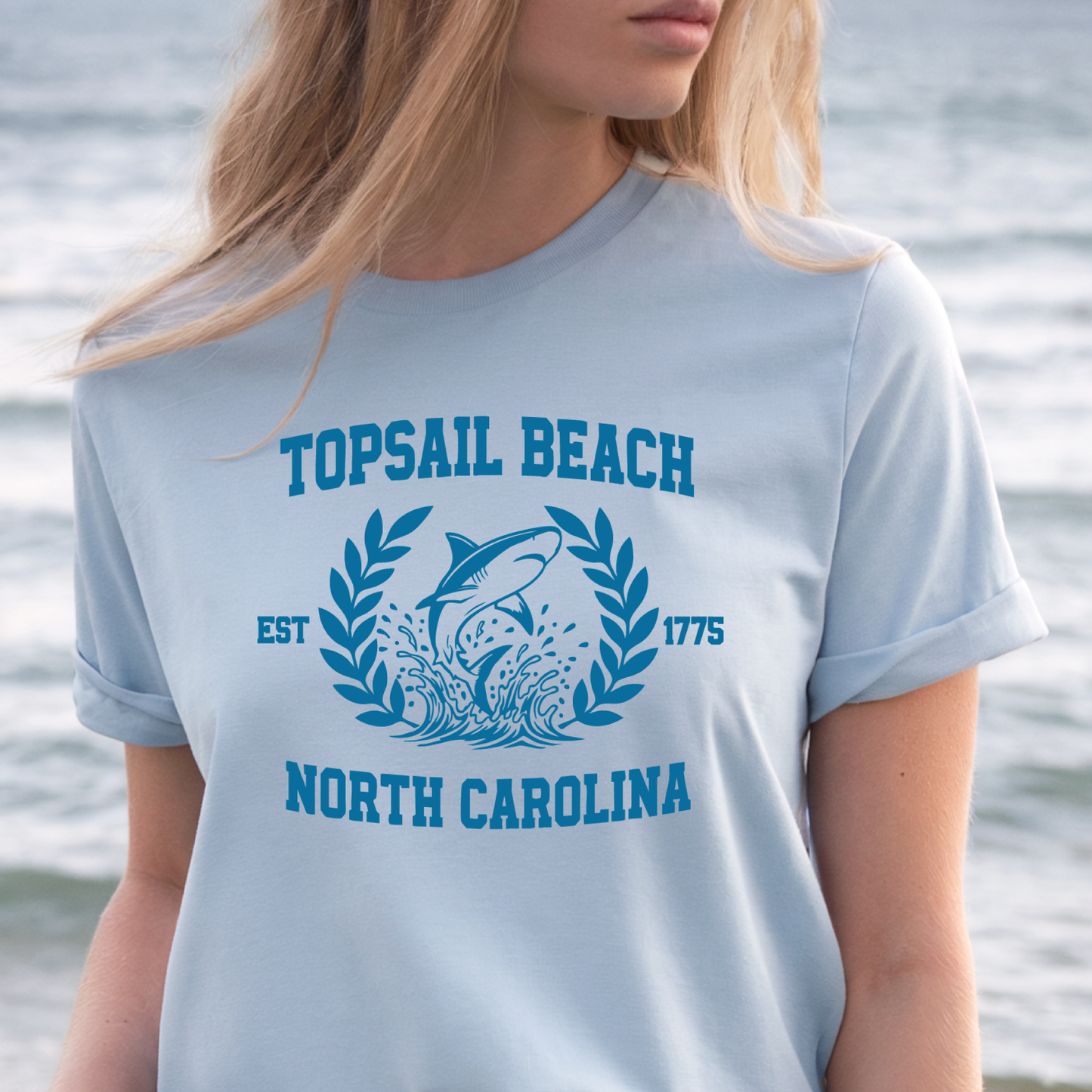 Person wearing a light blue t-shirt with 'Topical Beach, North Carolina' design by the water.