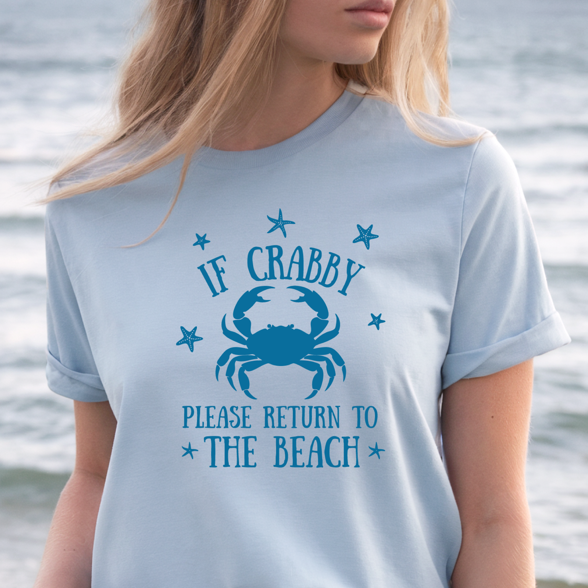 Person wearing a light blue t-shirt with a crab graphic and text by the water.