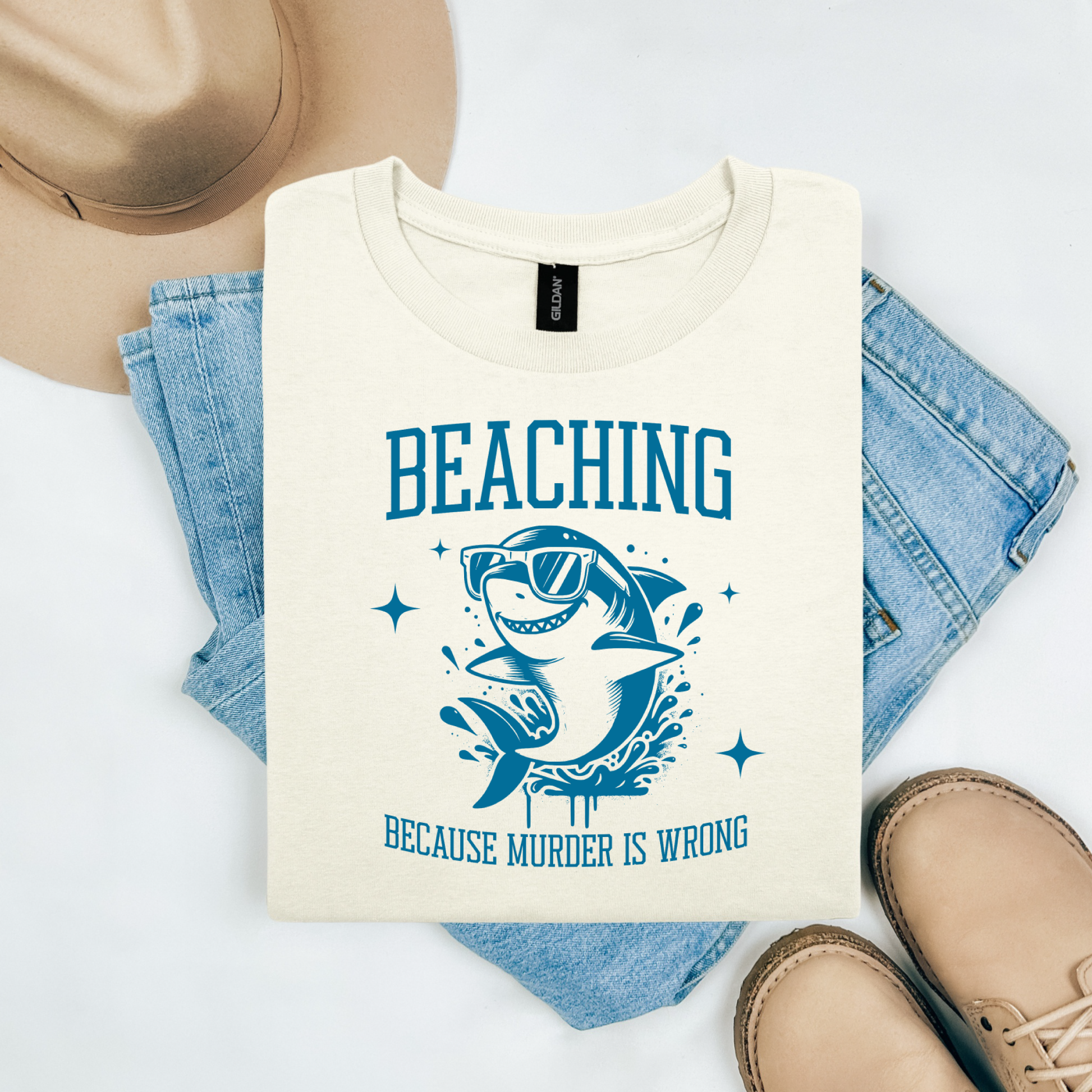Gildan 5000 "Beaching Because Murder Is Wrong" t shirt with a funny shark with sunglasses jumping out of water in one color graphics. Picture on a flay lay mockup with pair of blue jeans, brown shoes and a hat accessory.