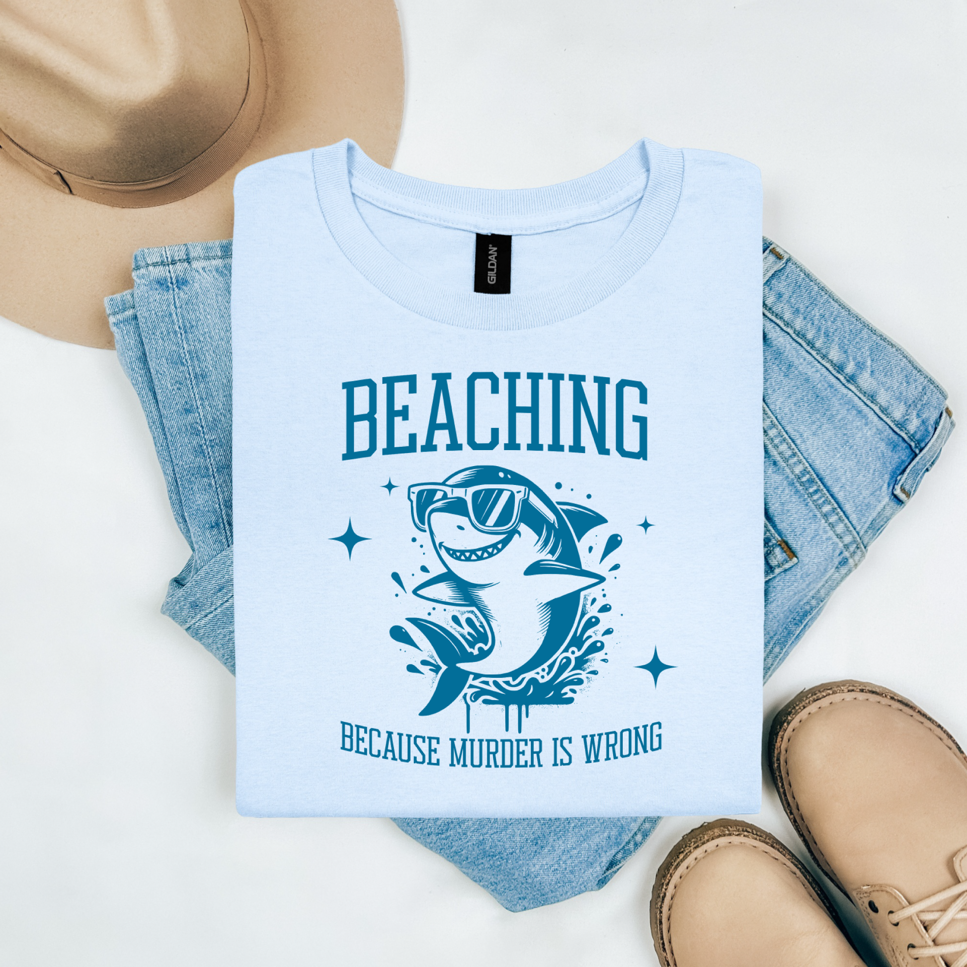 Gildan 5000 "Beaching Because Murder Is Wrong" t shirt with a funny shark with sunglasses jumping out of water in one color graphics. Picture on a flay lay mockup with pair of blue jeans, brown shoes and a hat accessory.