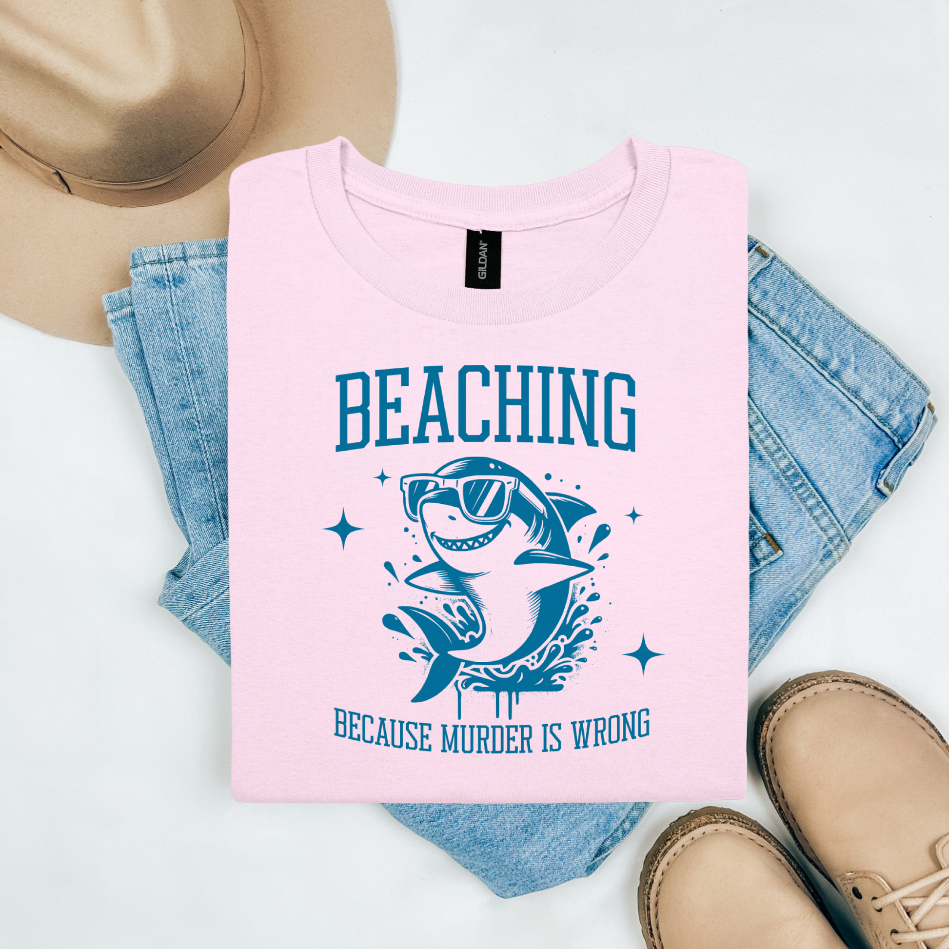 Gildan 5000 "Beaching Because Murder Is Wrong" t shirt with a funny shark with sunglasses jumping out of water in one color graphics. Picture on a flay lay mockup with pair of blue jeans, brown shoes and a hat accessory.