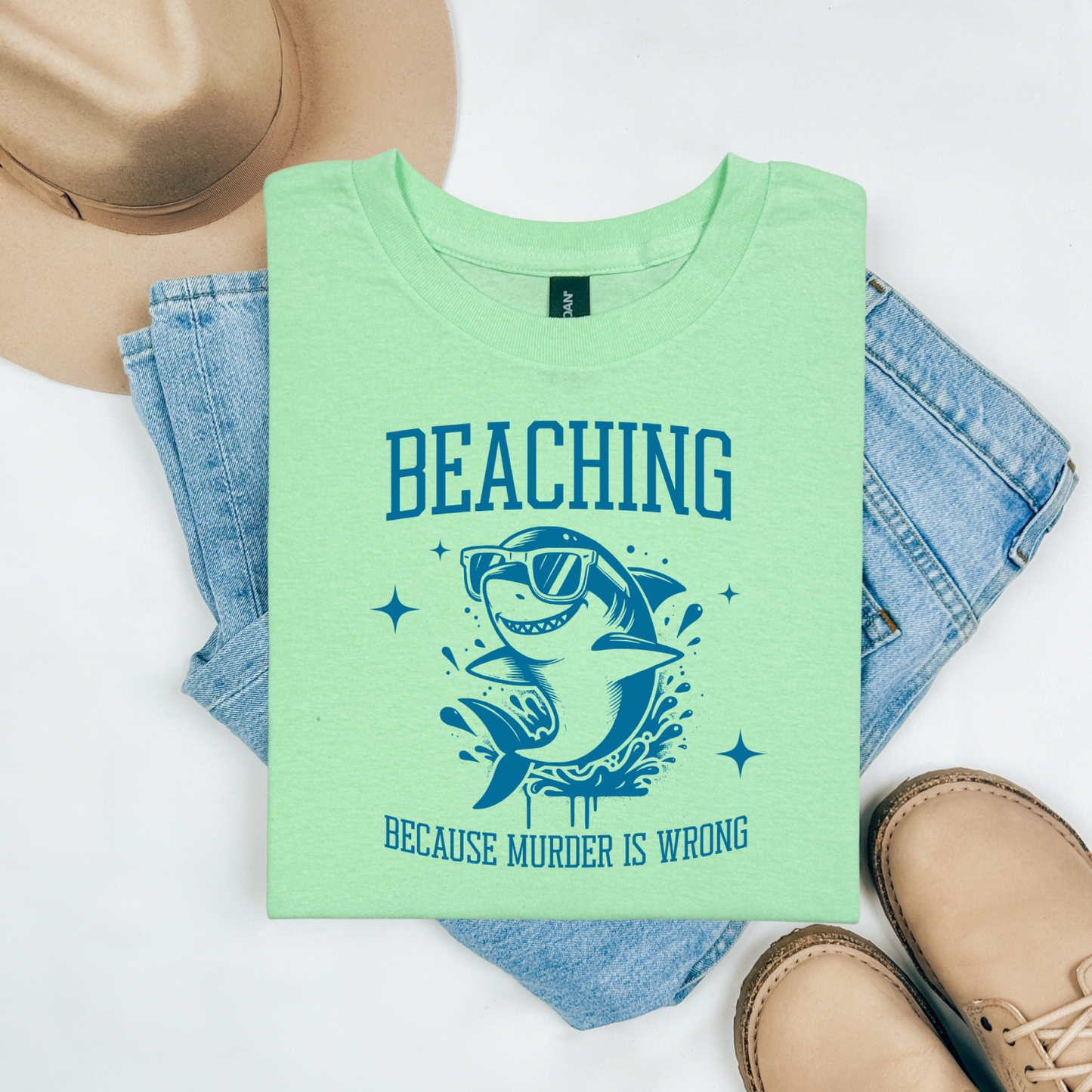 Gildan 5000 "Beaching Because Murder Is Wrong" t shirt with a funny shark with sunglasses jumping out of water in one color graphics. Picture on a flay lay mockup with pair of blue jeans, brown shoes and a hat accessory.
