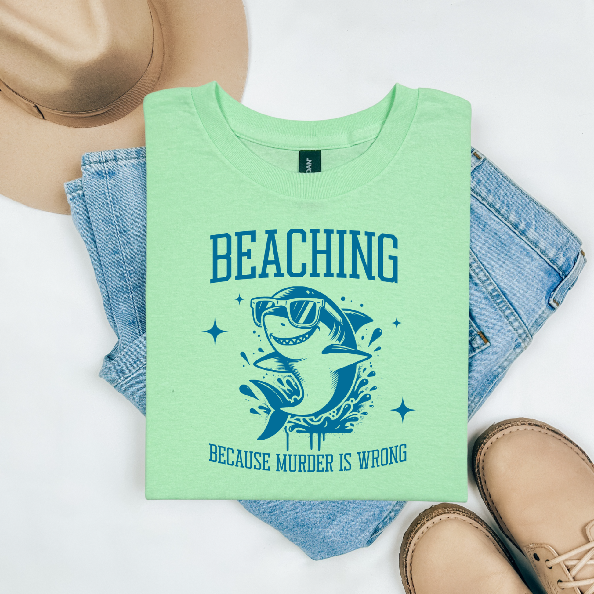 Gildan 5000 "Beaching Because Murder Is Wrong" t shirt with a funny shark with sunglasses jumping out of water in one color graphics. Picture on a flay lay mockup with pair of blue jeans, brown shoes and a hat accessory.