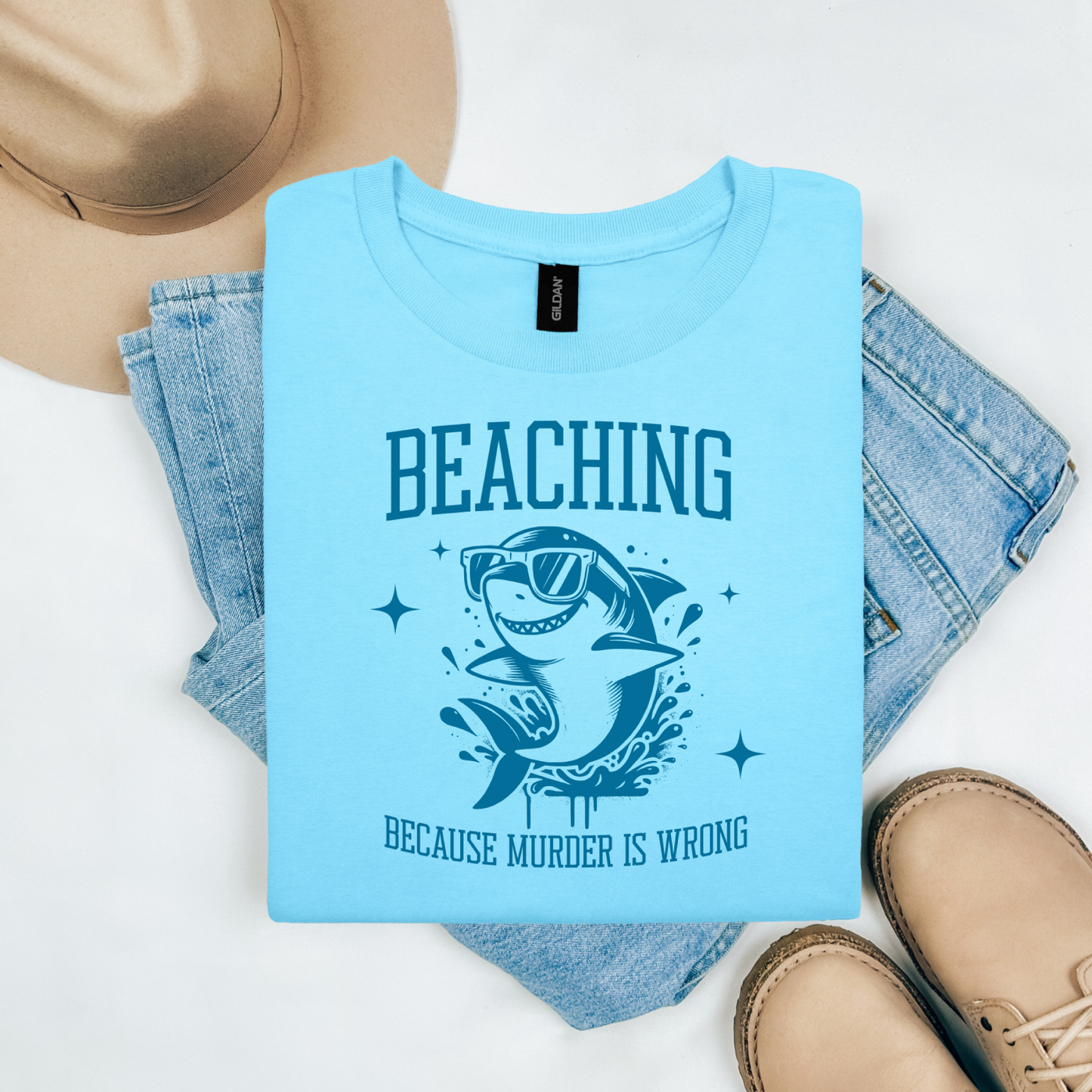 Gildan 5000 "Beaching Because Murder Is Wrong" t shirt with a funny shark with sunglasses jumping out of water in one color graphics. Picture on a flay lay mockup with pair of blue jeans, brown shoes and a hat accessory.