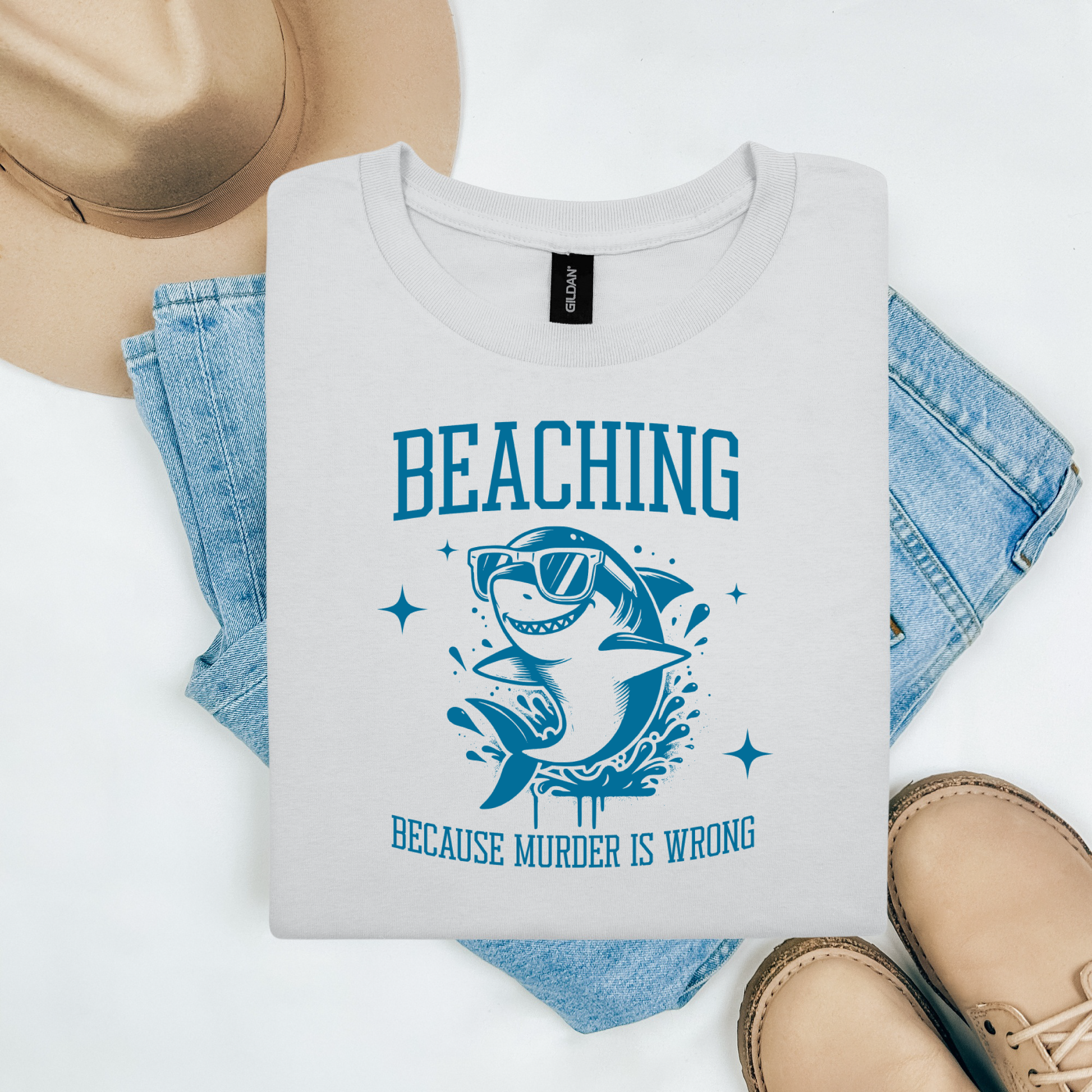 Gildan 5000 "Beaching Because Murder Is Wrong" t shirt with a funny shark with sunglasses jumping out of water in one color graphics. Picture on a flay lay mockup with pair of blue jeans, brown shoes and a hat accessory.