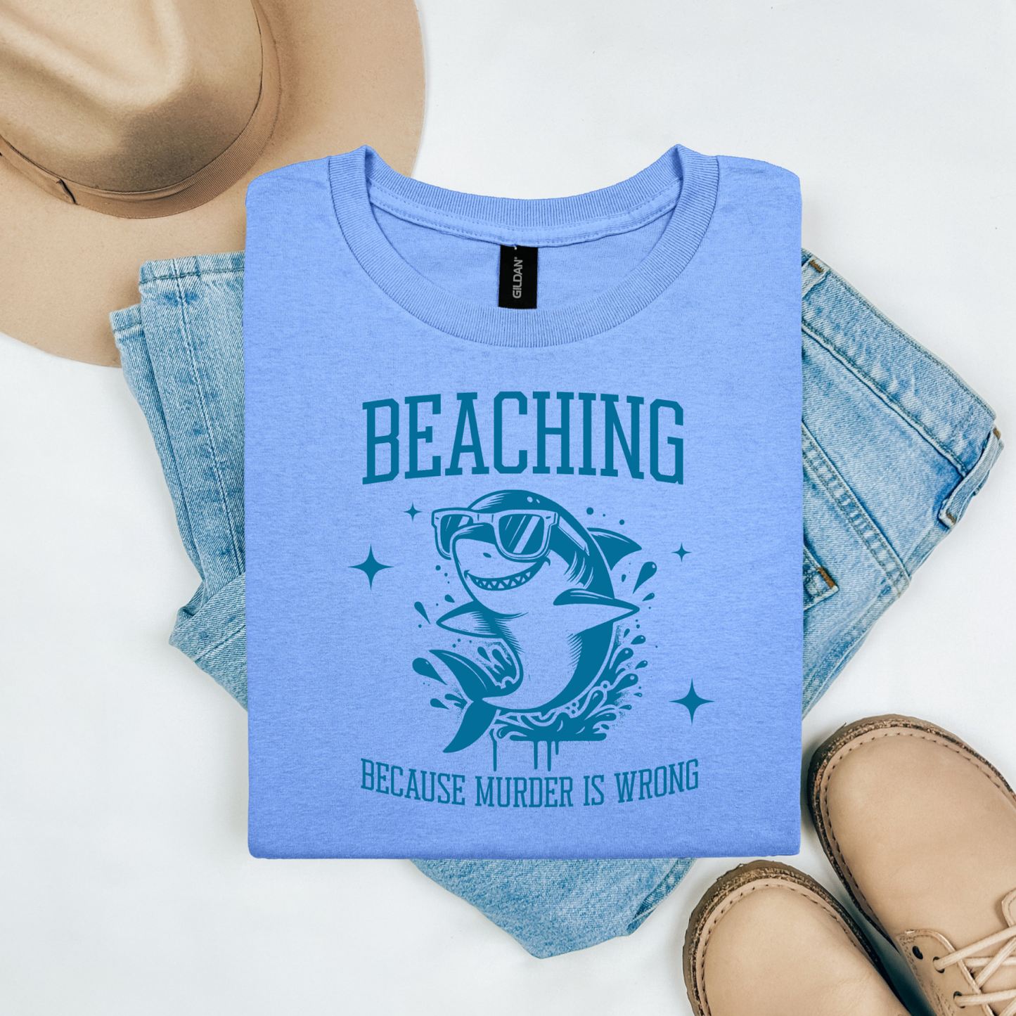 Gildan 5000 "Beaching Because Murder Is Wrong" t shirt with a funny shark with sunglasses jumping out of water in one color graphics. Picture on a flay lay mockup with pair of blue jeans, brown shoes and a hat accessory.