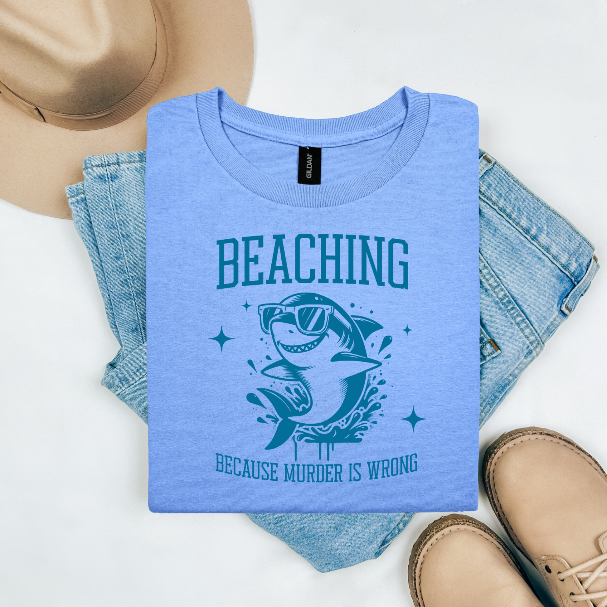 Gildan 5000 "Beaching Because Murder Is Wrong" t shirt with a funny shark with sunglasses jumping out of water in one color graphics. Picture on a flay lay mockup with pair of blue jeans, brown shoes and a hat accessory.