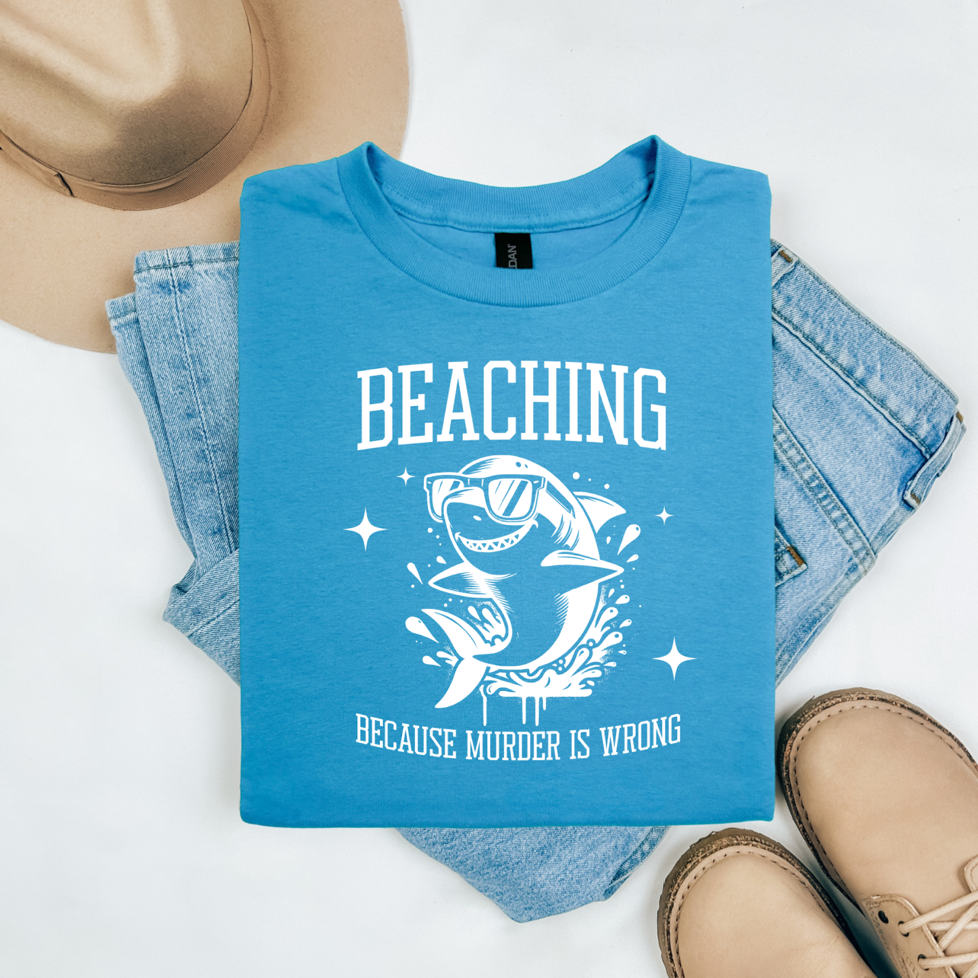 Gildan 5000 "Beaching Because Murder Is Wrong" t shirt with a funny shark with sunglasses jumping out of water in one color graphics. Picture on a flay lay mockup with pair of blue jeans, brown shoes and a hat accessory.