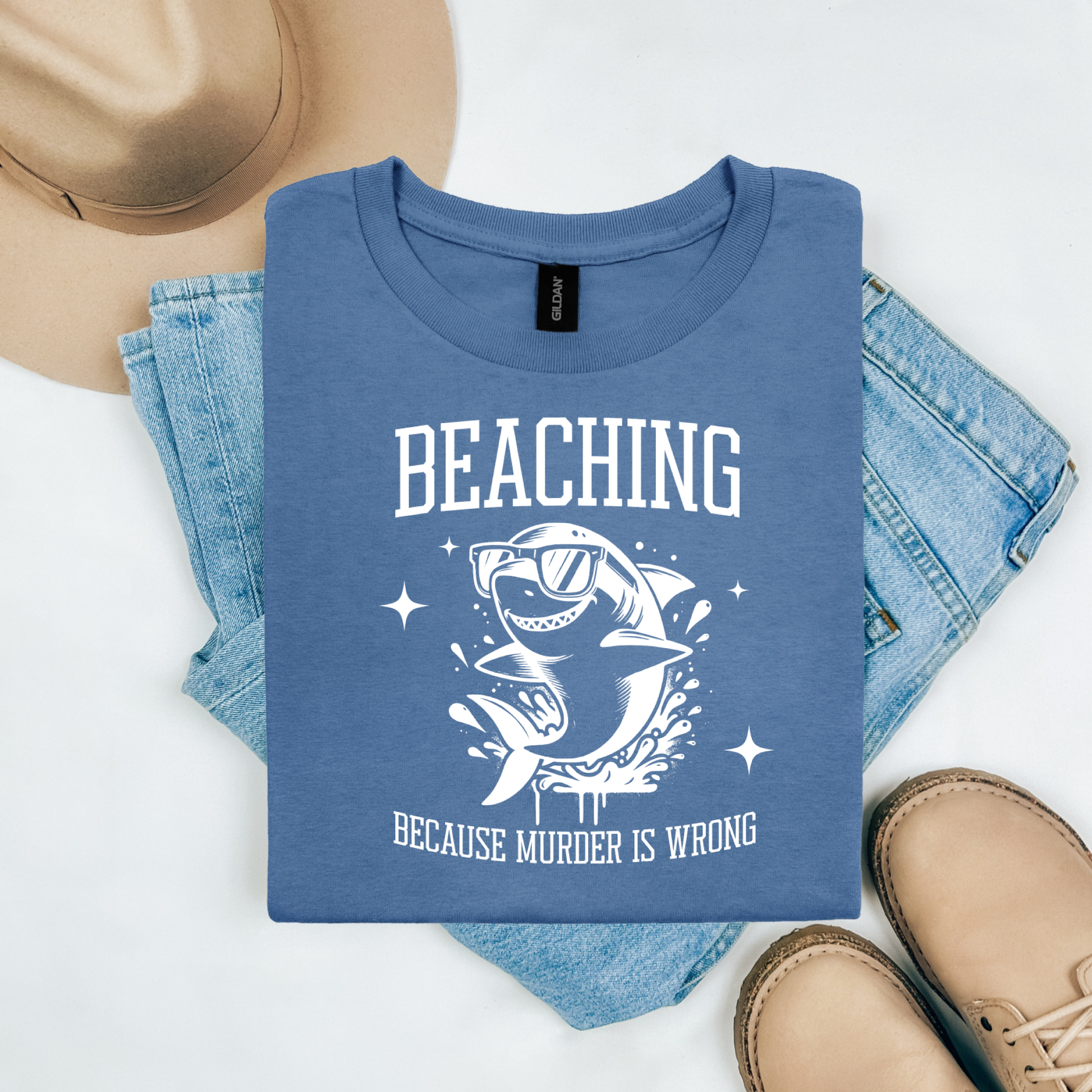 Gildan 5000 "Beaching Because Murder Is Wrong" t shirt with a funny shark with sunglasses jumping out of water in one color graphics. Picture on a flay lay mockup with pair of blue jeans, brown shoes and a hat accessory.