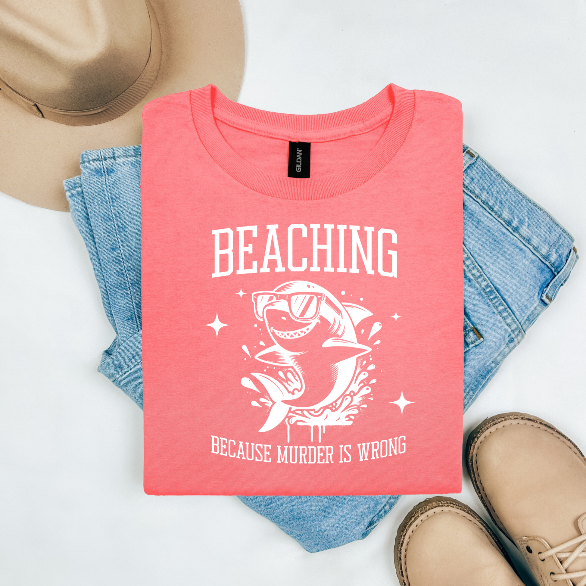 Gildan 5000 "Beaching Because Murder Is Wrong" t shirt with a funny shark with sunglasses jumping out of water in one color graphics. Picture on a flay lay mockup with pair of blue jeans, brown shoes and a hat accessory.