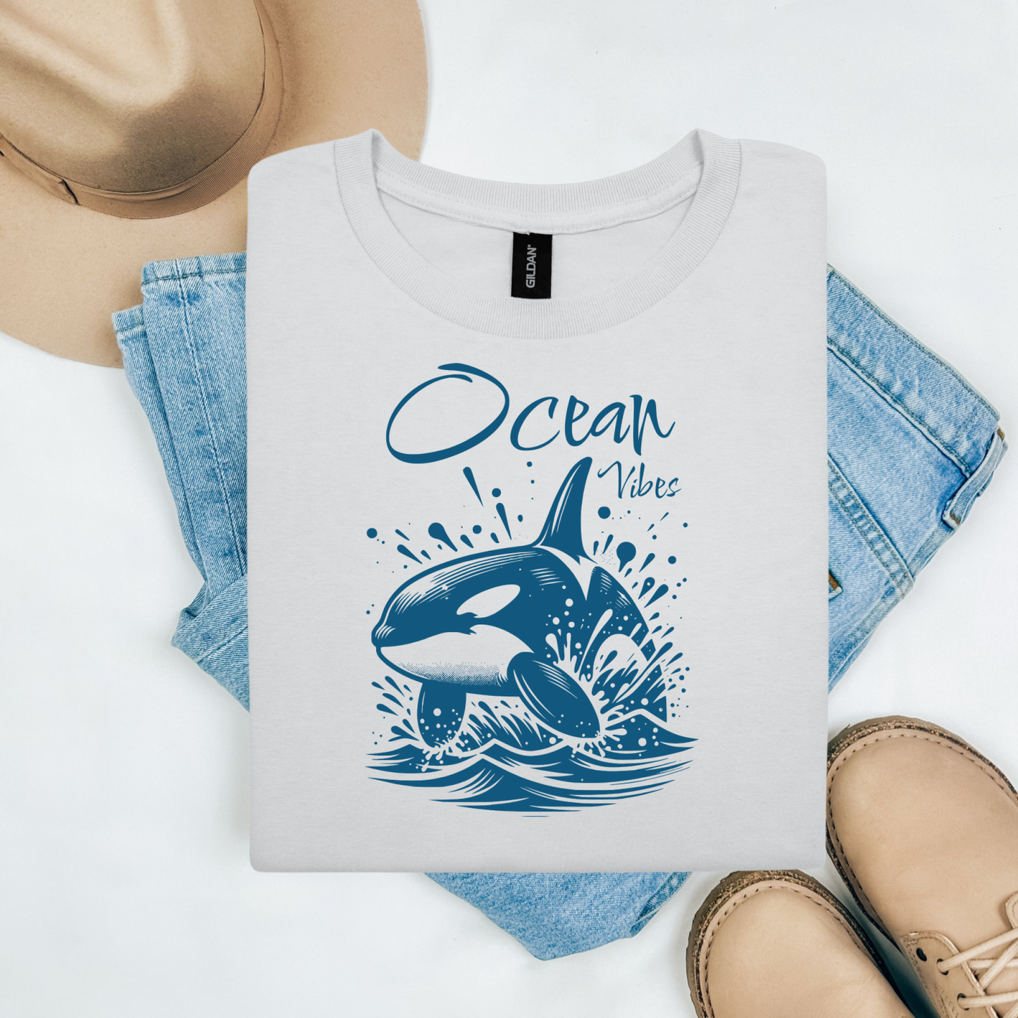 Gildan 5000 "Ocean Vibes" t shirt with a orca whale jumping out of water graphics in a one color graphic. Picture on a flay lay mockup with pair of blue jeans, brown shoes and a hat accessory.