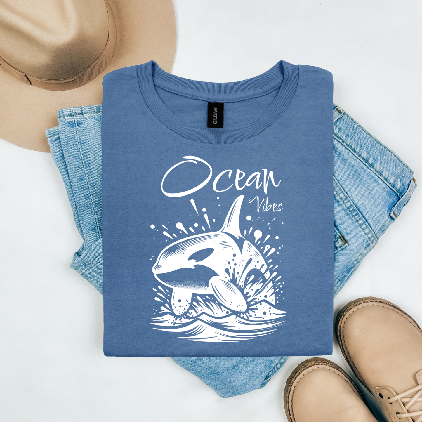 Gildan 5000 "Ocean Vibes" t shirt with a orca whale jumping out of water graphics in a one color graphic. Picture on a flay lay mockup with pair of blue jeans, brown shoes and a hat accessory.