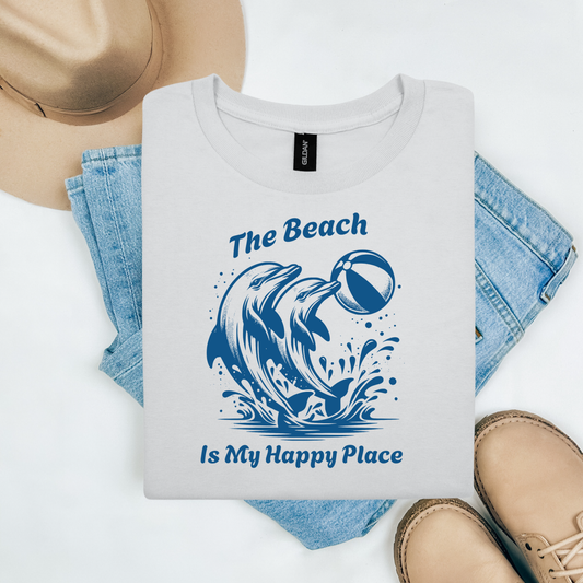 Gildan 5000 "The Beach is my Happy Place" t shirt with dolphins playing with beach ball jumping out of ocean graphics in monochrome color. Picture on a flay lay mockup with pair of blue jeans, brown shoes and a hat accessory.