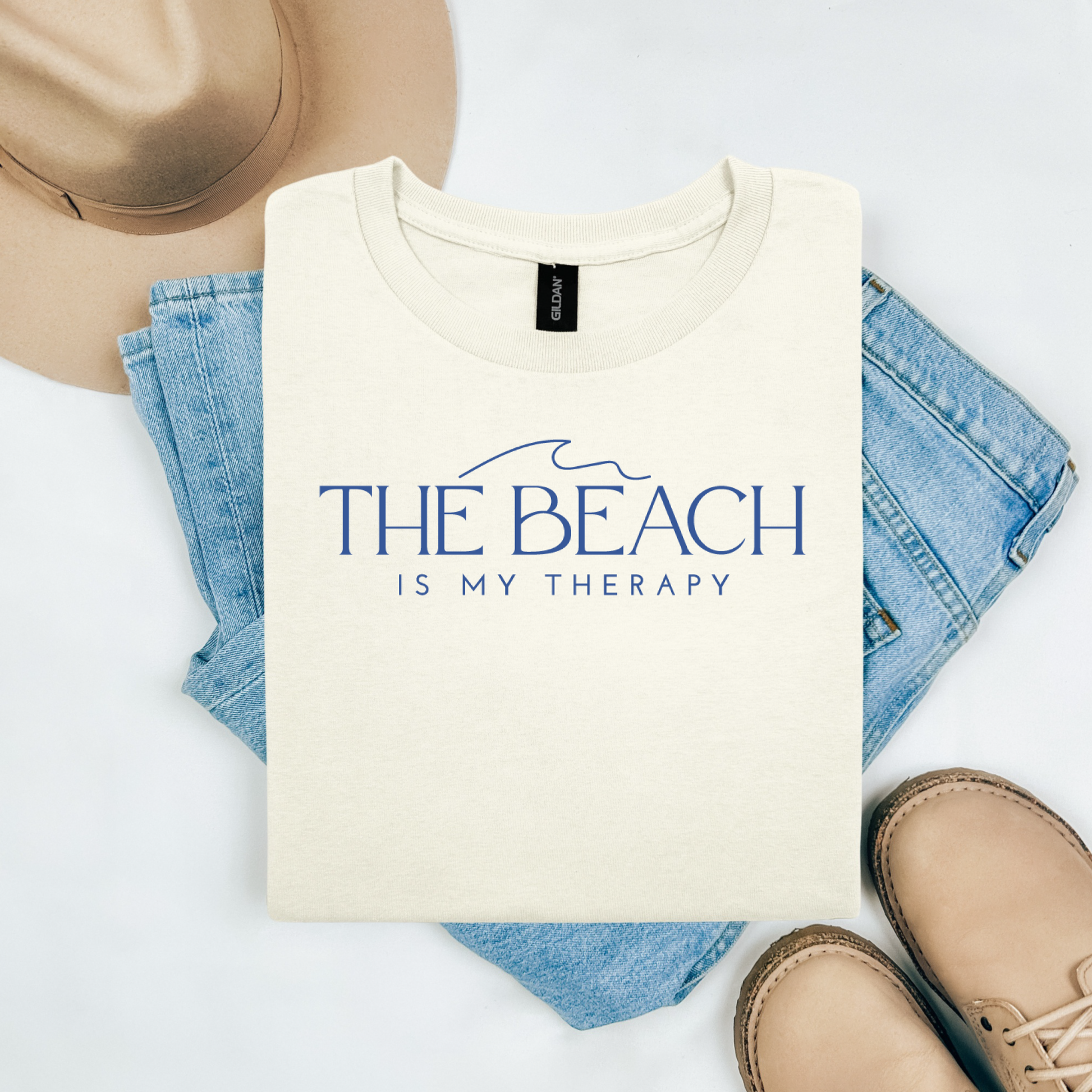 Gildan 5000 "The Beach Is My Therapy" t shirt with a simple wave graphic in one color. Picture on a flay lay mockup with pair of blue jeans, brown shoes and a hat accessory.