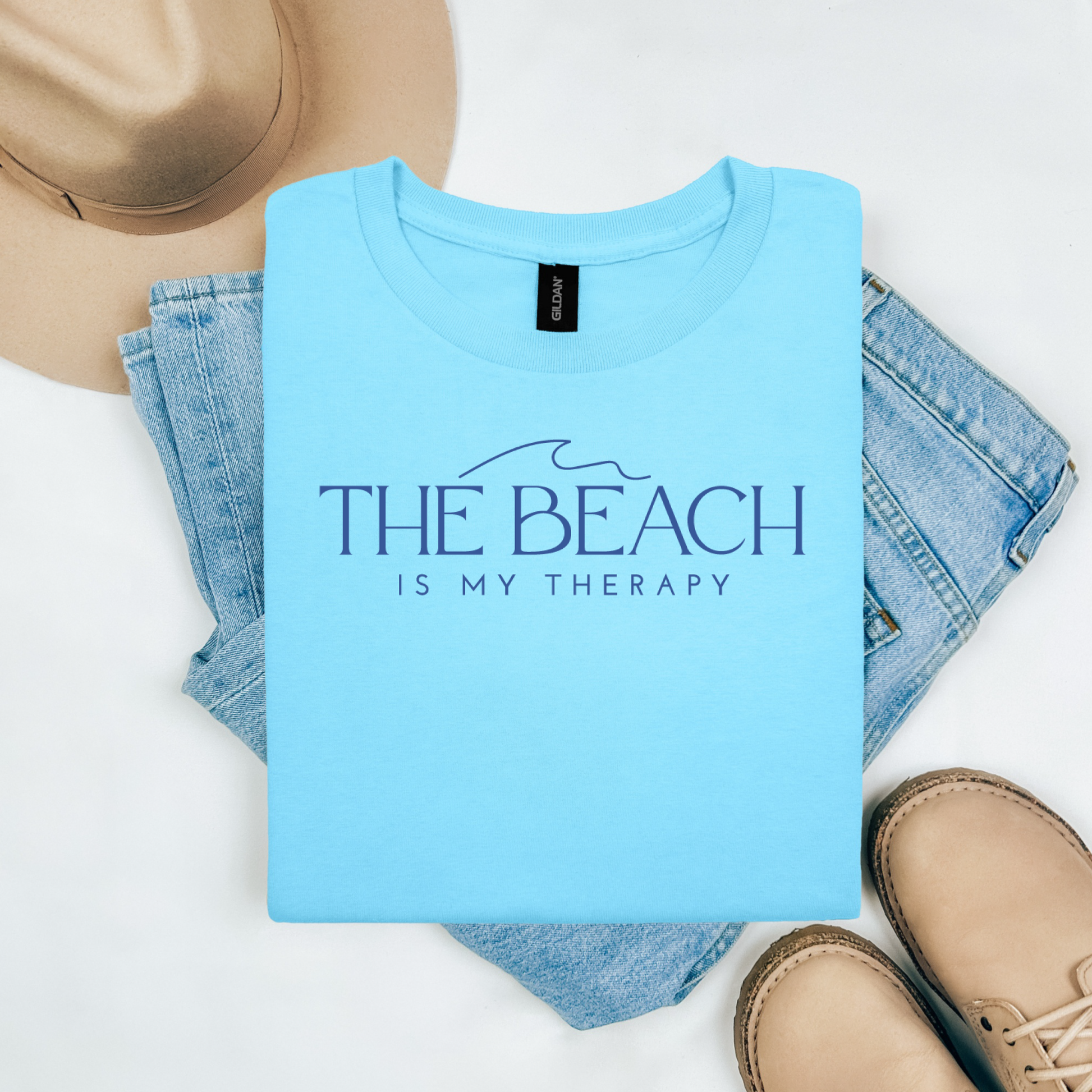Gildan 5000 "The Beach Is My Therapy" t shirt with a simple wave graphic in one color. Picture on a flay lay mockup with pair of blue jeans, brown shoes and a hat accessory.