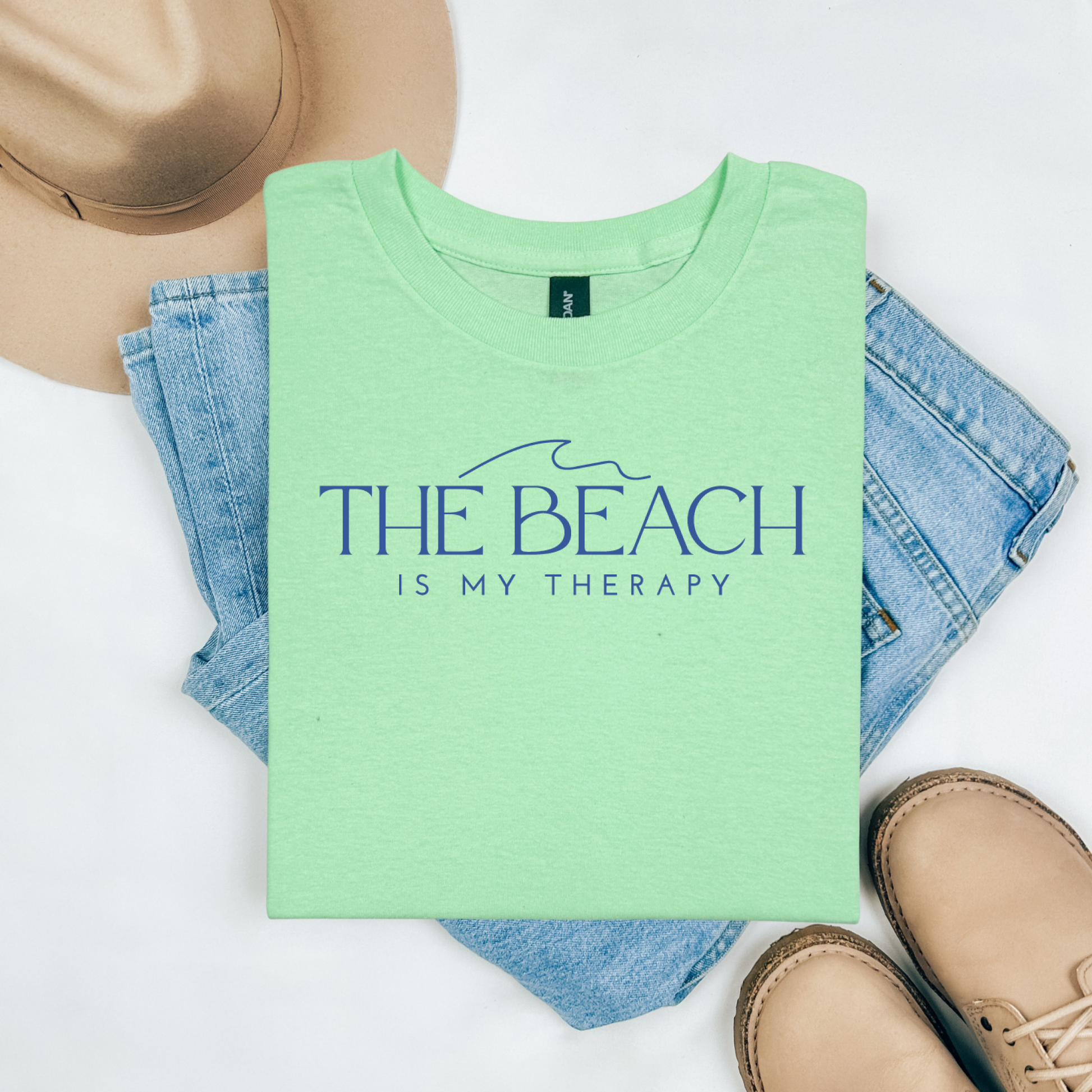 Gildan 5000 "The Beach Is My Therapy" t shirt with a simple wave graphic in one color. Picture on a flay lay mockup with pair of blue jeans, brown shoes and a hat accessory.