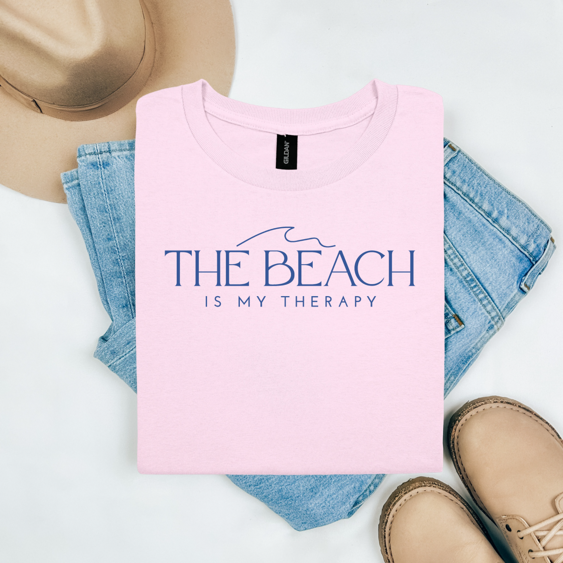 Gildan 5000 "The Beach Is My Therapy" t shirt with a simple wave graphic in one color. Picture on a flay lay mockup with pair of blue jeans, brown shoes and a hat accessory.
