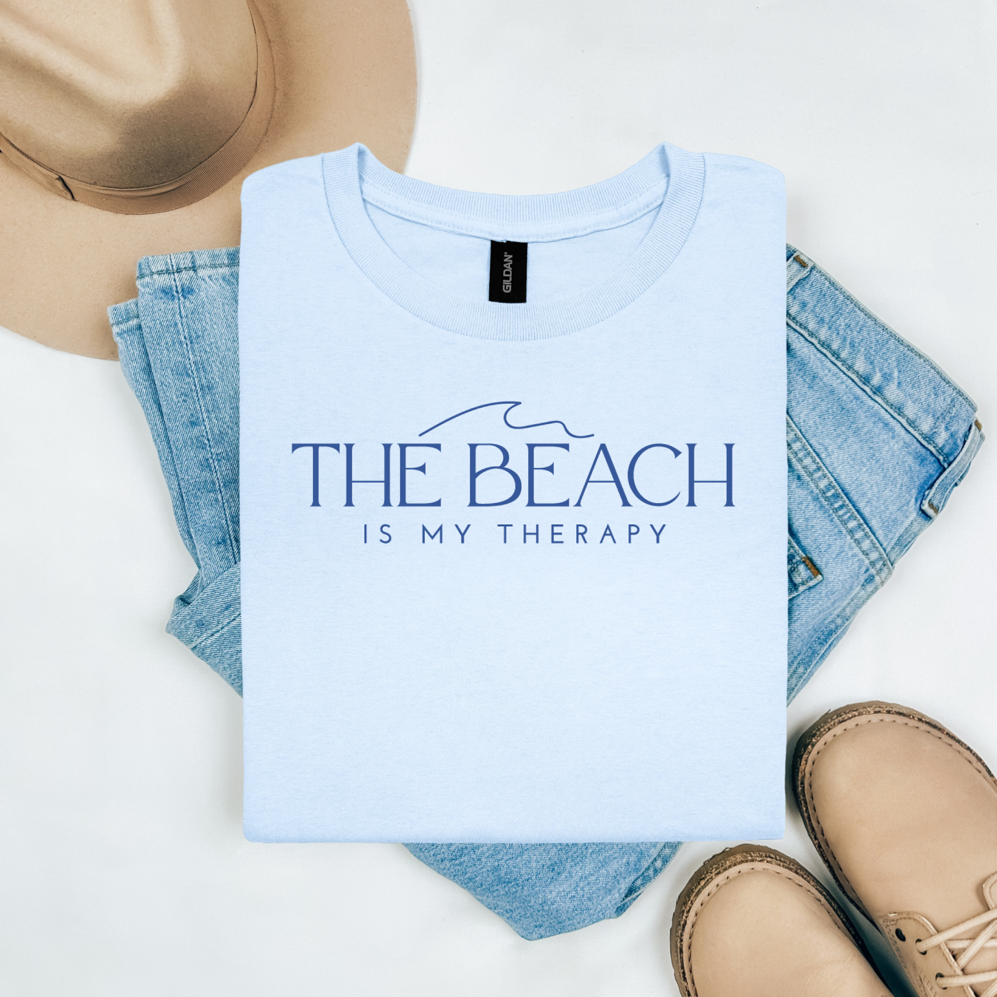 Gildan 5000 "The Beach Is My Therapy" t shirt with a simple wave graphic in one color. Picture on a flay lay mockup with pair of blue jeans, brown shoes and a hat accessory.