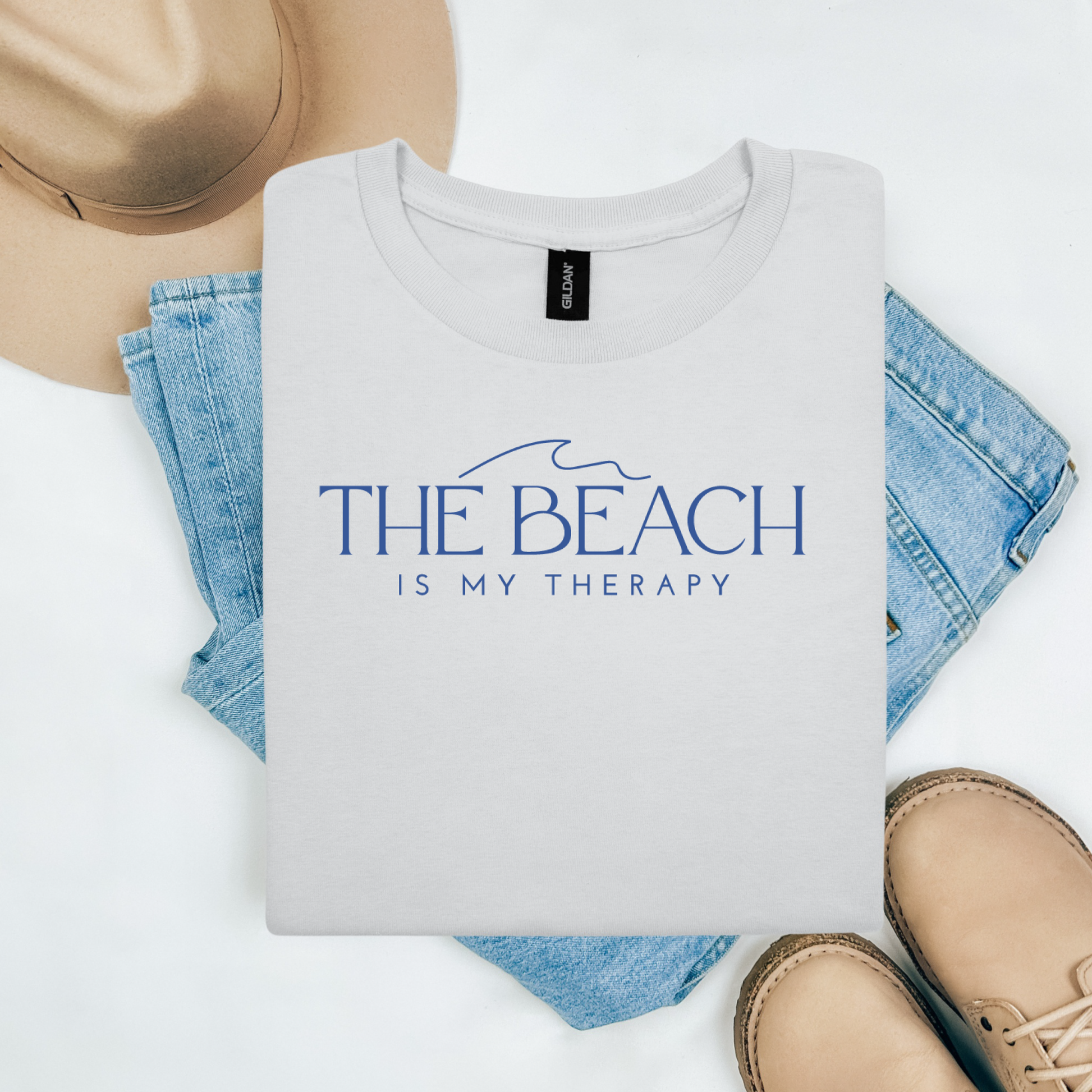 Gildan 5000 "The Beach Is My Therapy" t shirt with a simple wave graphic in one color. Picture on a flay lay mockup with pair of blue jeans, brown shoes and a hat accessory.