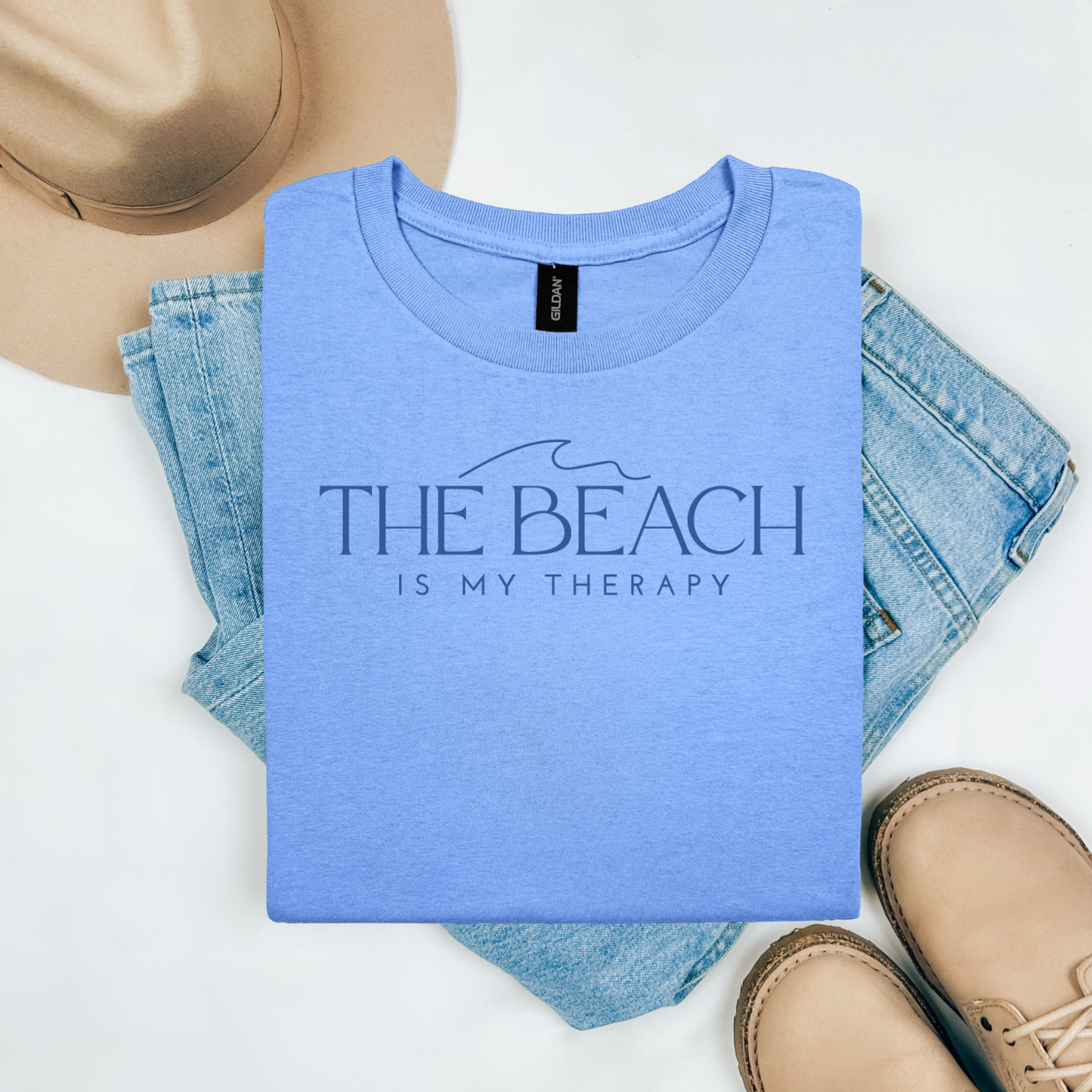 Gildan 5000 "The Beach Is My Therapy" t shirt with a simple wave graphic in one color. Picture on a flay lay mockup with pair of blue jeans, brown shoes and a hat accessory.