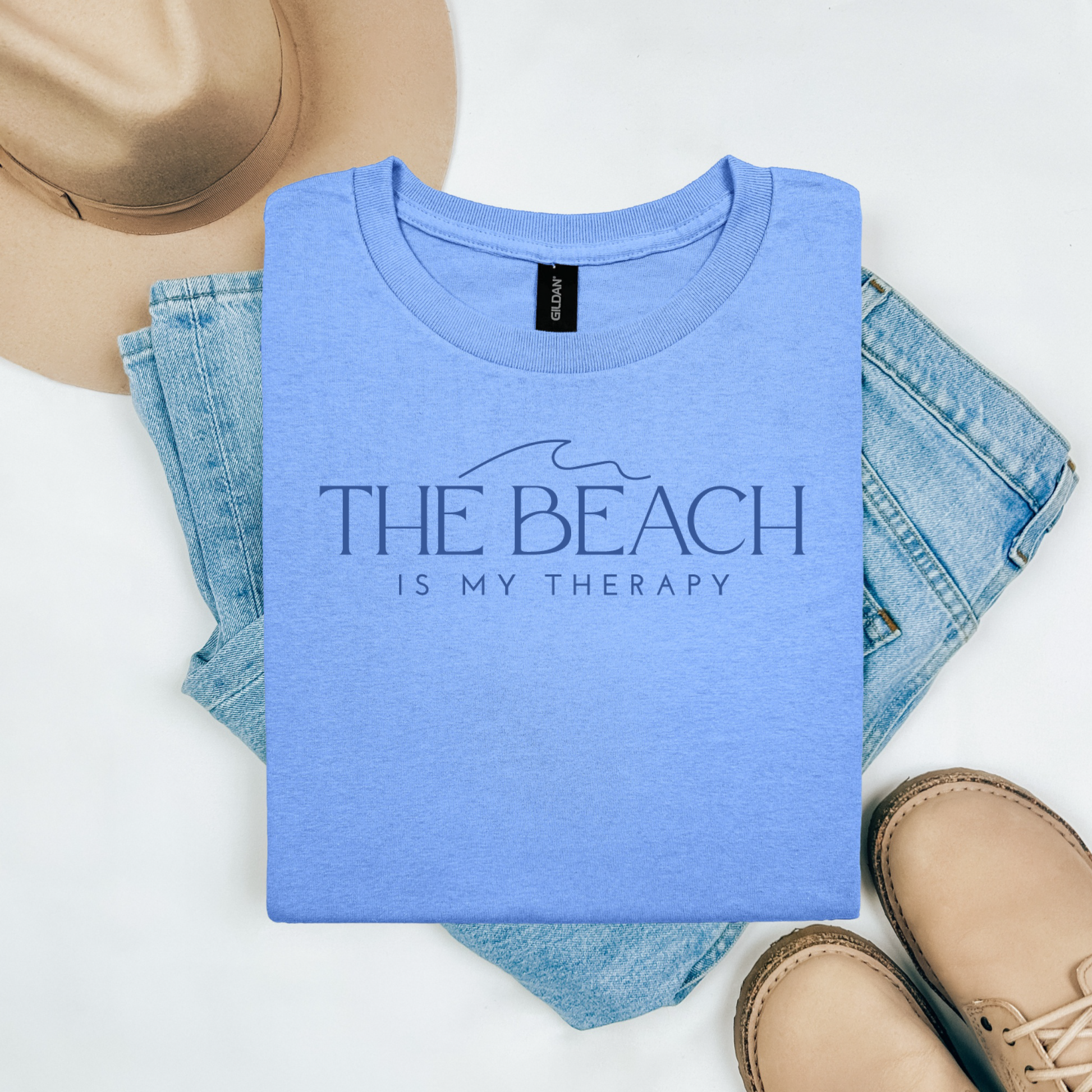 Gildan 5000 "The Beach Is My Therapy" t shirt with a simple wave graphic in one color. Picture on a flay lay mockup with pair of blue jeans, brown shoes and a hat accessory.